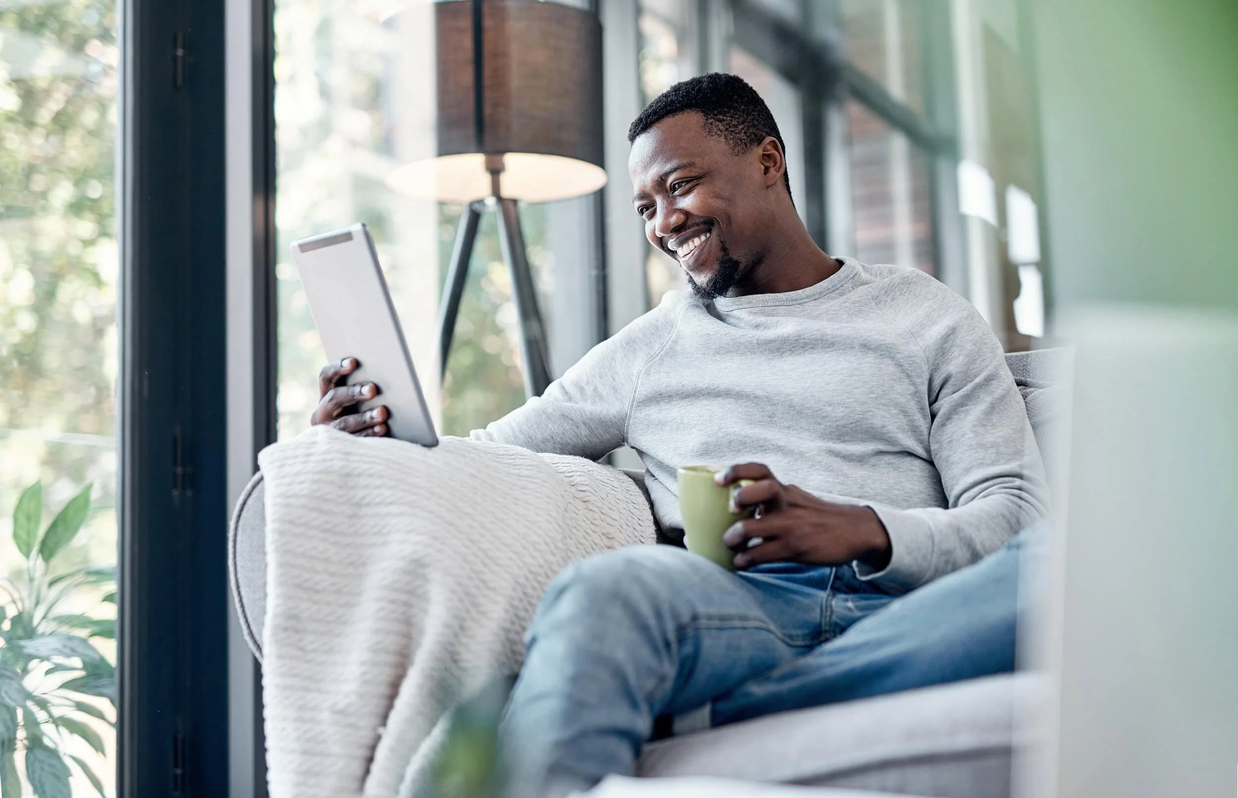 A smiling man sitting on a sofa, looking at a tablet, holding a yellow mug, with a window and a lamp in the background.