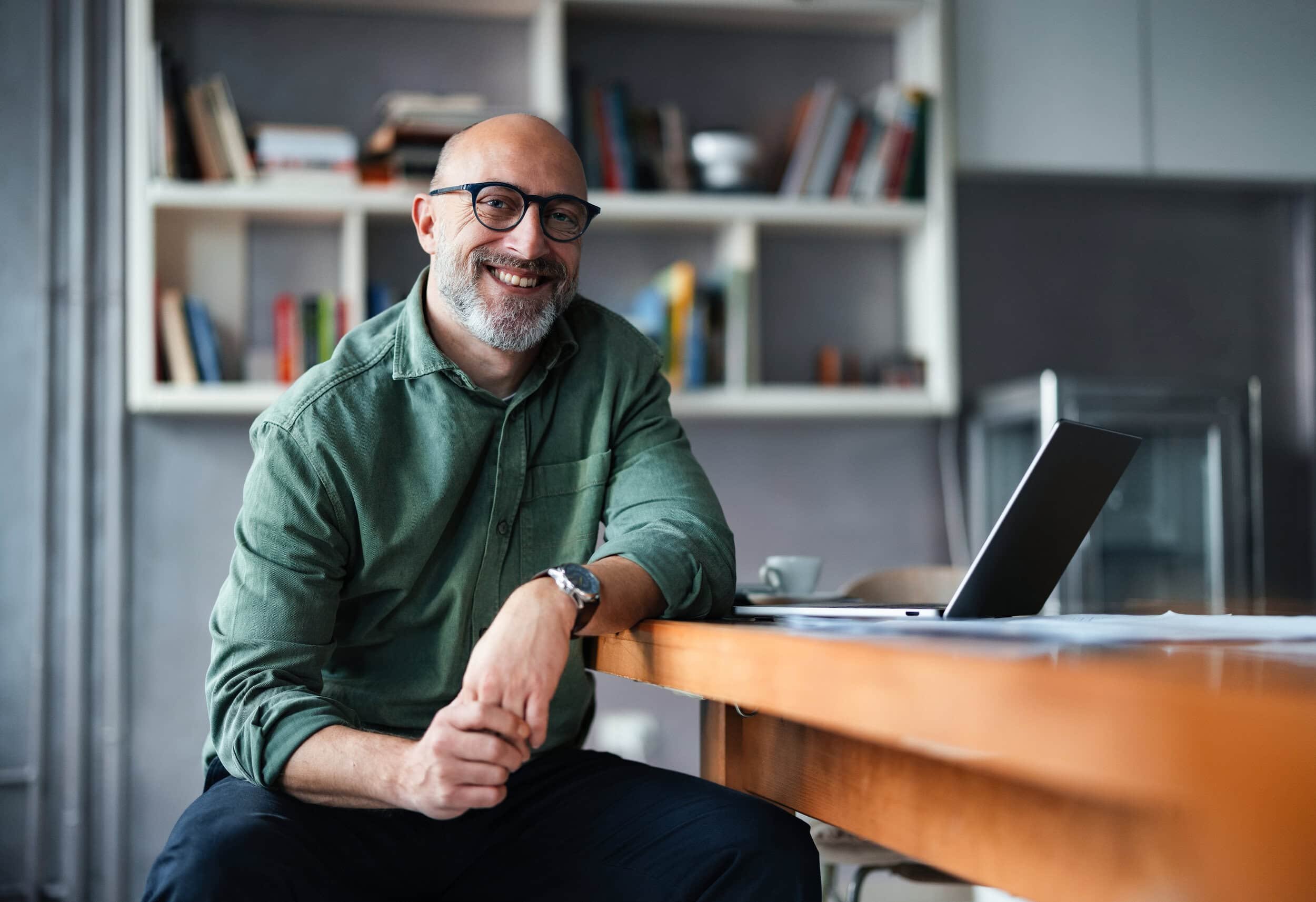 A smiling man with glasses and a beard wearing a green shirt, sitting at a desk with a laptop in a room with bookshelves.
