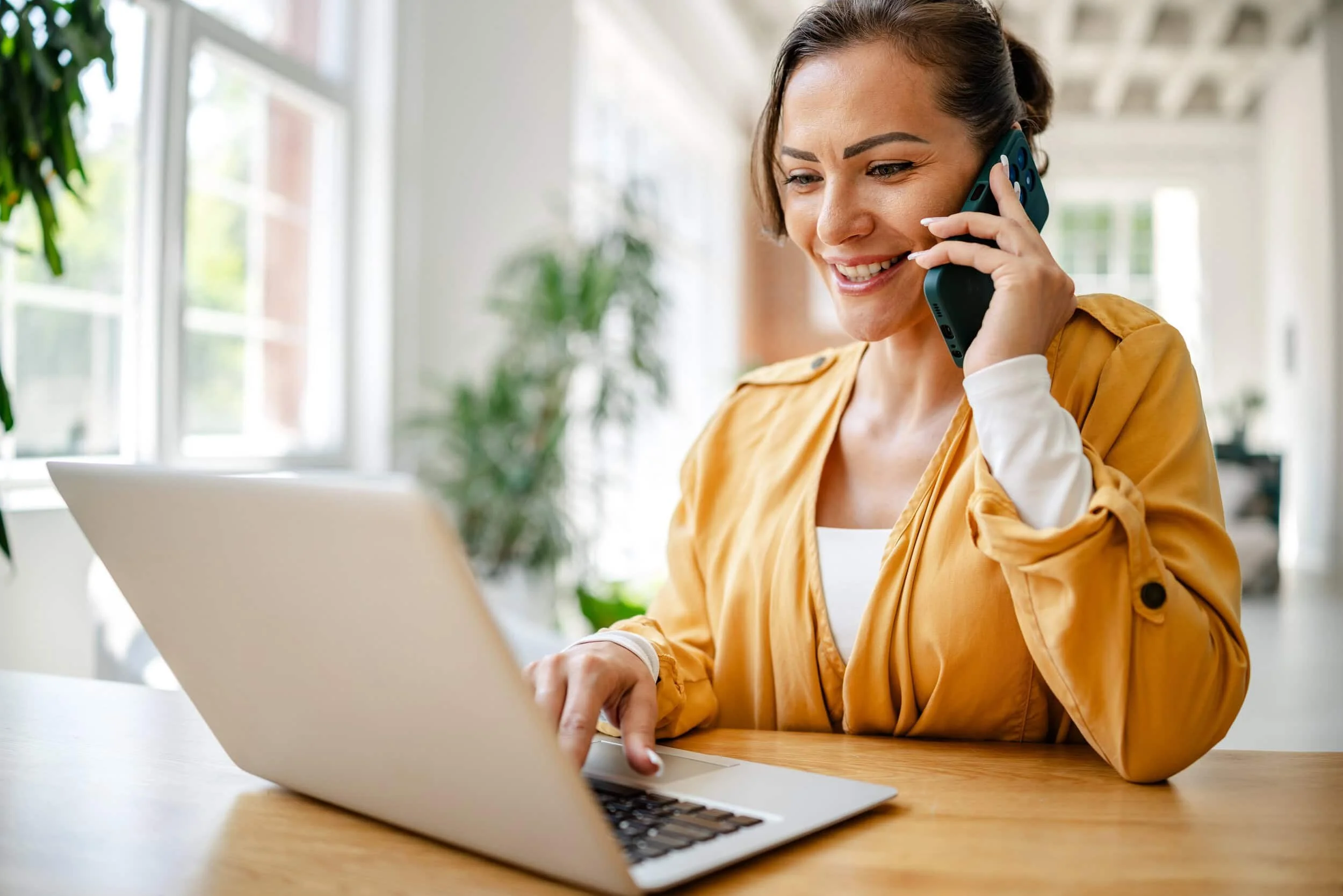 Woman smiling while talking on the phone and using a laptop in a bright, modern room with plants.