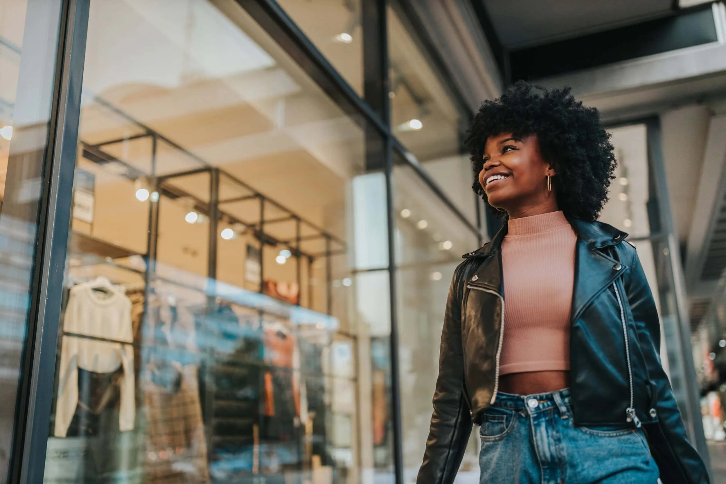 Young woman with curly hair smiling and walking past a store window in an urban shopping district.