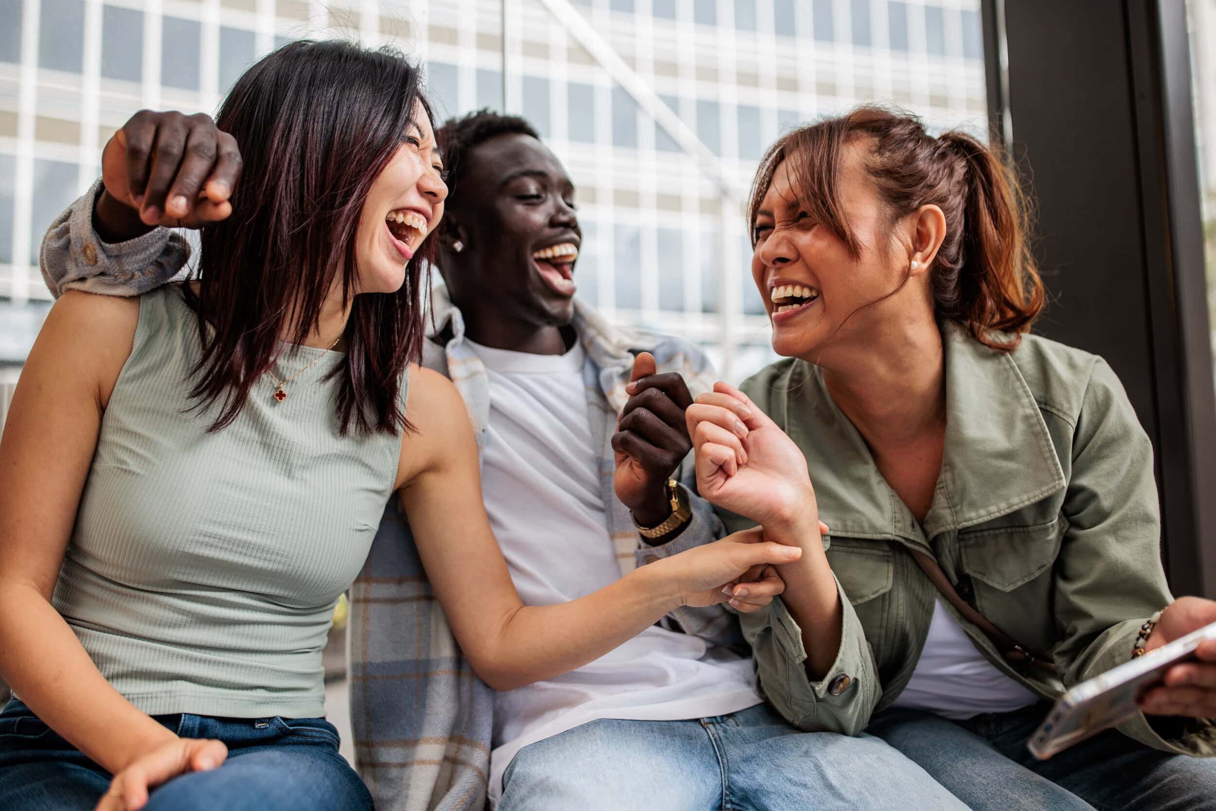 Three women laughing and smiling together indoors, sitting on a bench near a large window.