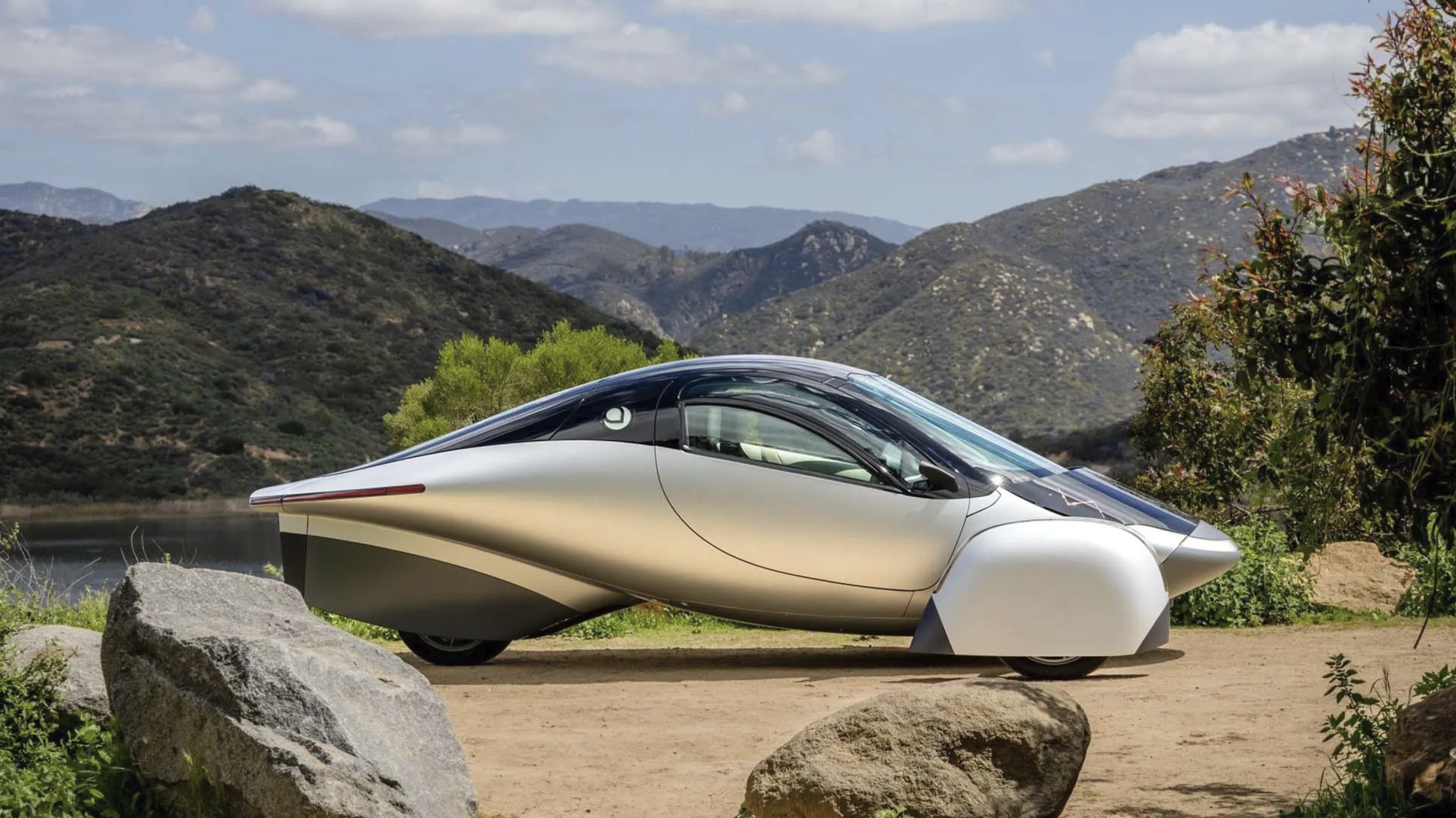 A futuristic silver and black luxury car with a sleek design, parked outdoors on a dirt surface with rocks, green plants, and a backdrop of mountains and a cloudy sky.