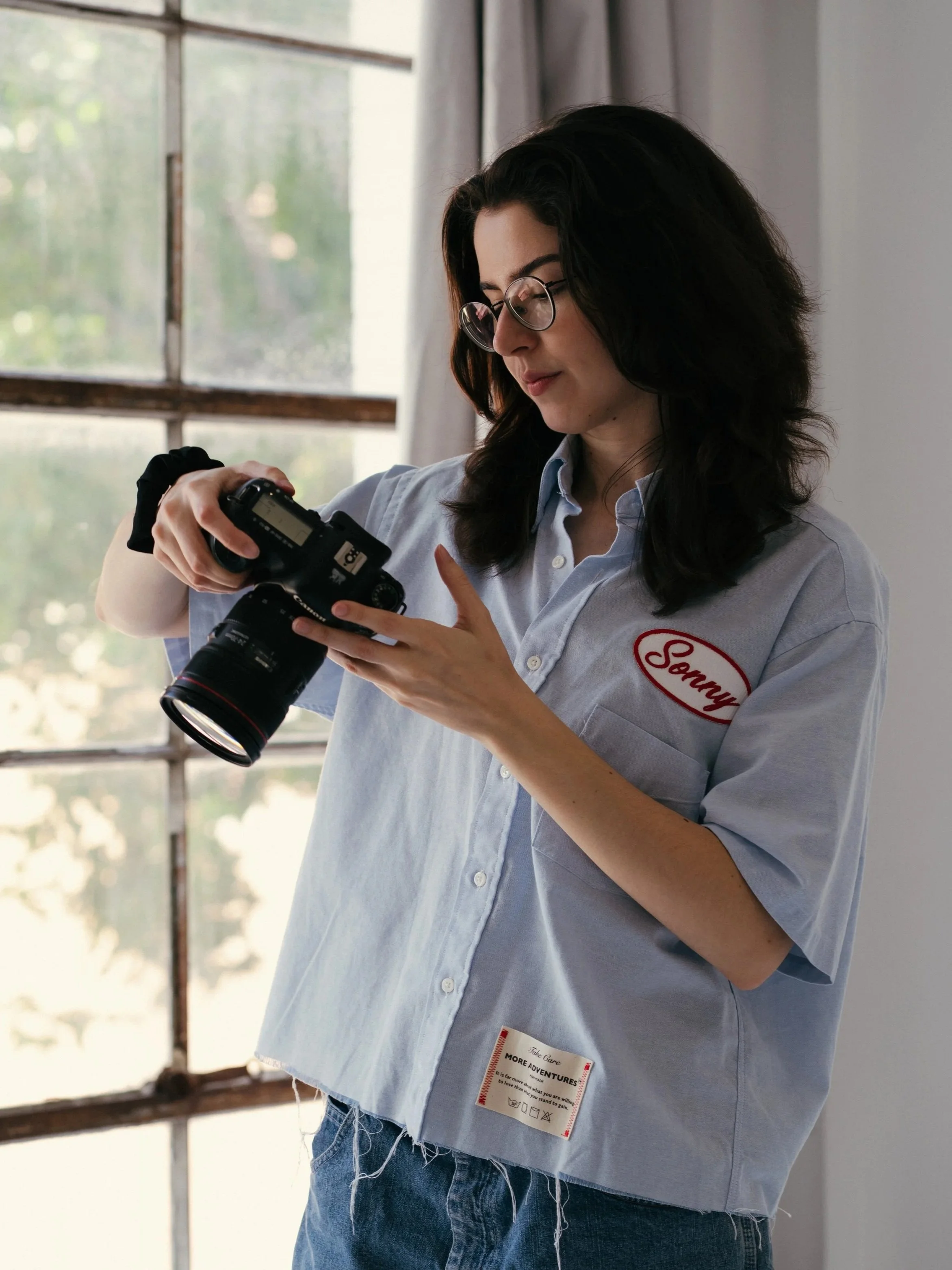 A woman with glasses and dark hair wearing a light blue button-up shirt looking at the screen of a camera she is holding.