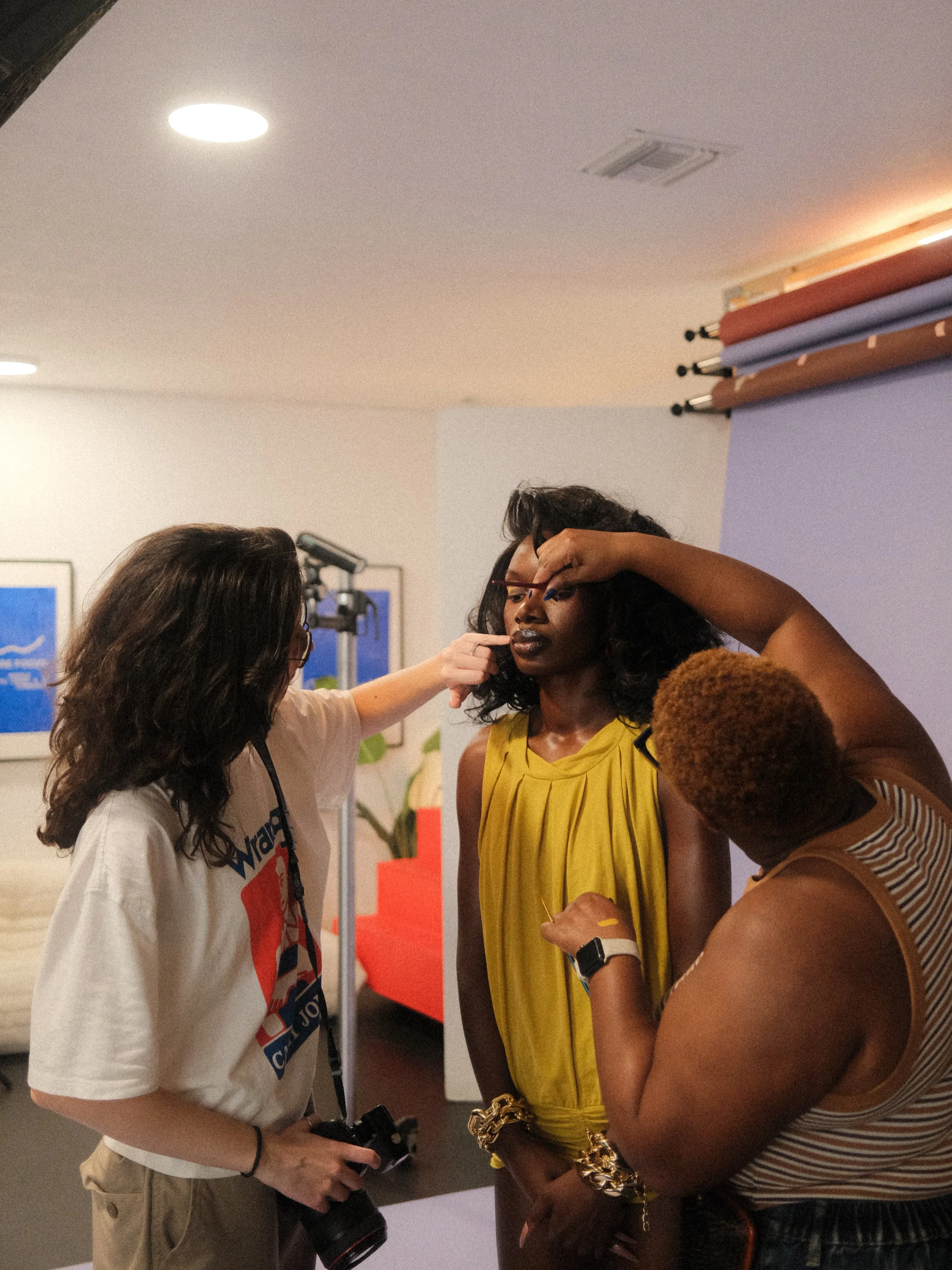 A woman in a yellow dress is having her makeup touched up by two makeup artists in a room with photography equipment and a purple backdrop.