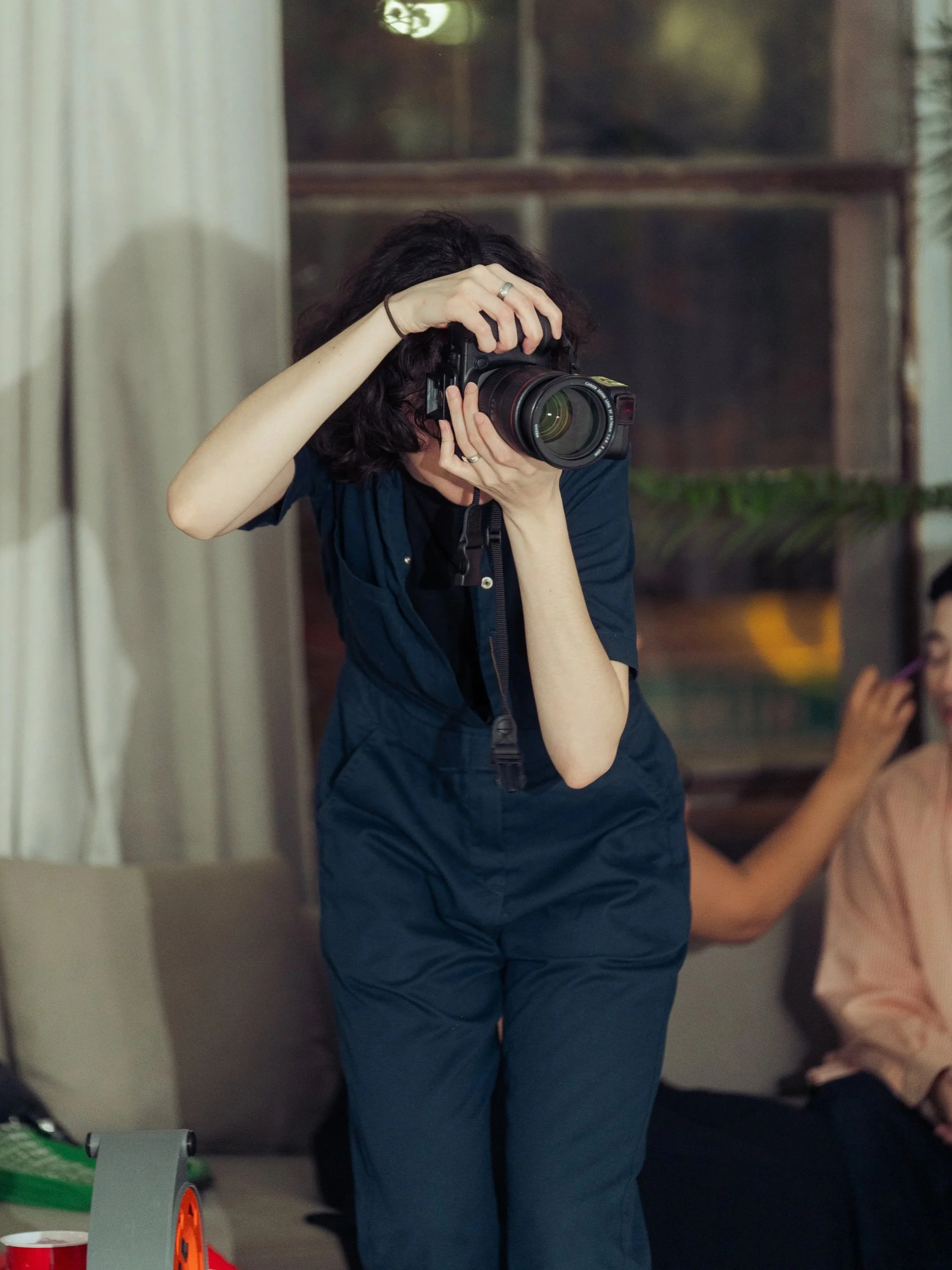 A woman is taking a photo with a professional camera while standing indoors, with a woman sitting on a sofa in the background.