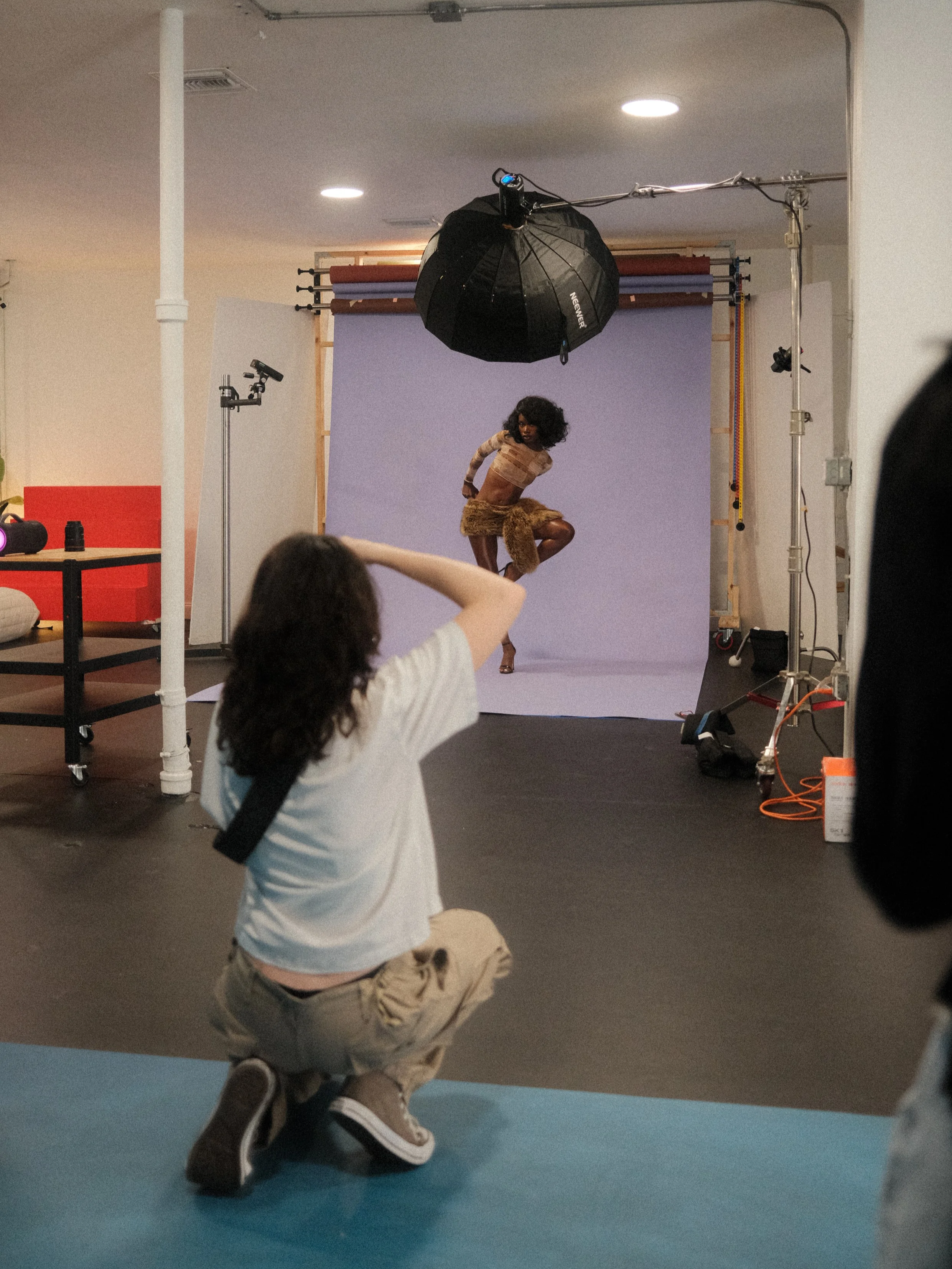 A photoshoot in a studio with a model dancing or posing, a photographer kneeling and taking pictures, and studio lighting and backdrop setup.