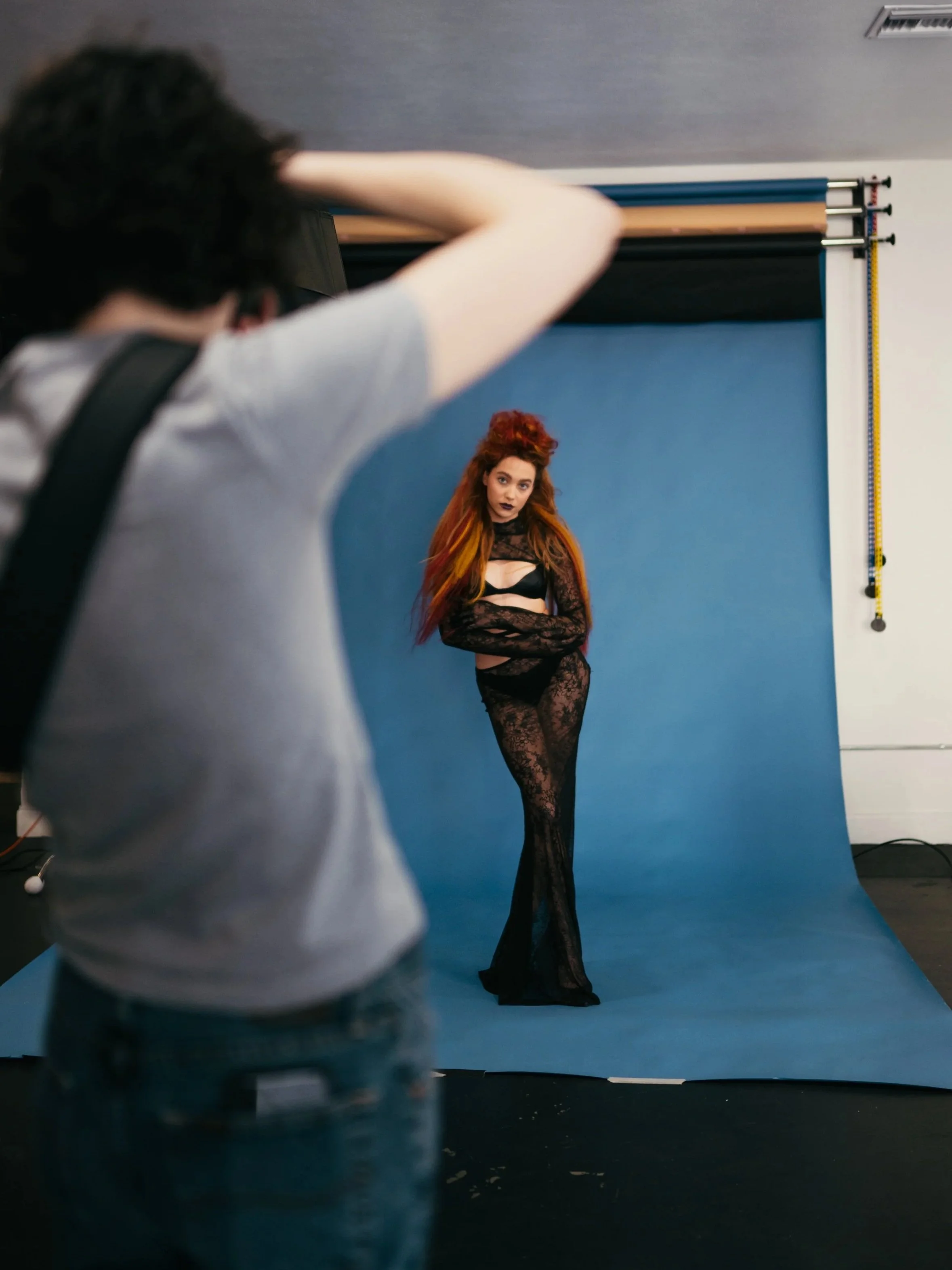 Photographer taking a picture of a woman with red hair and dark lipstick, posing against a blue backdrop in a photography studio.