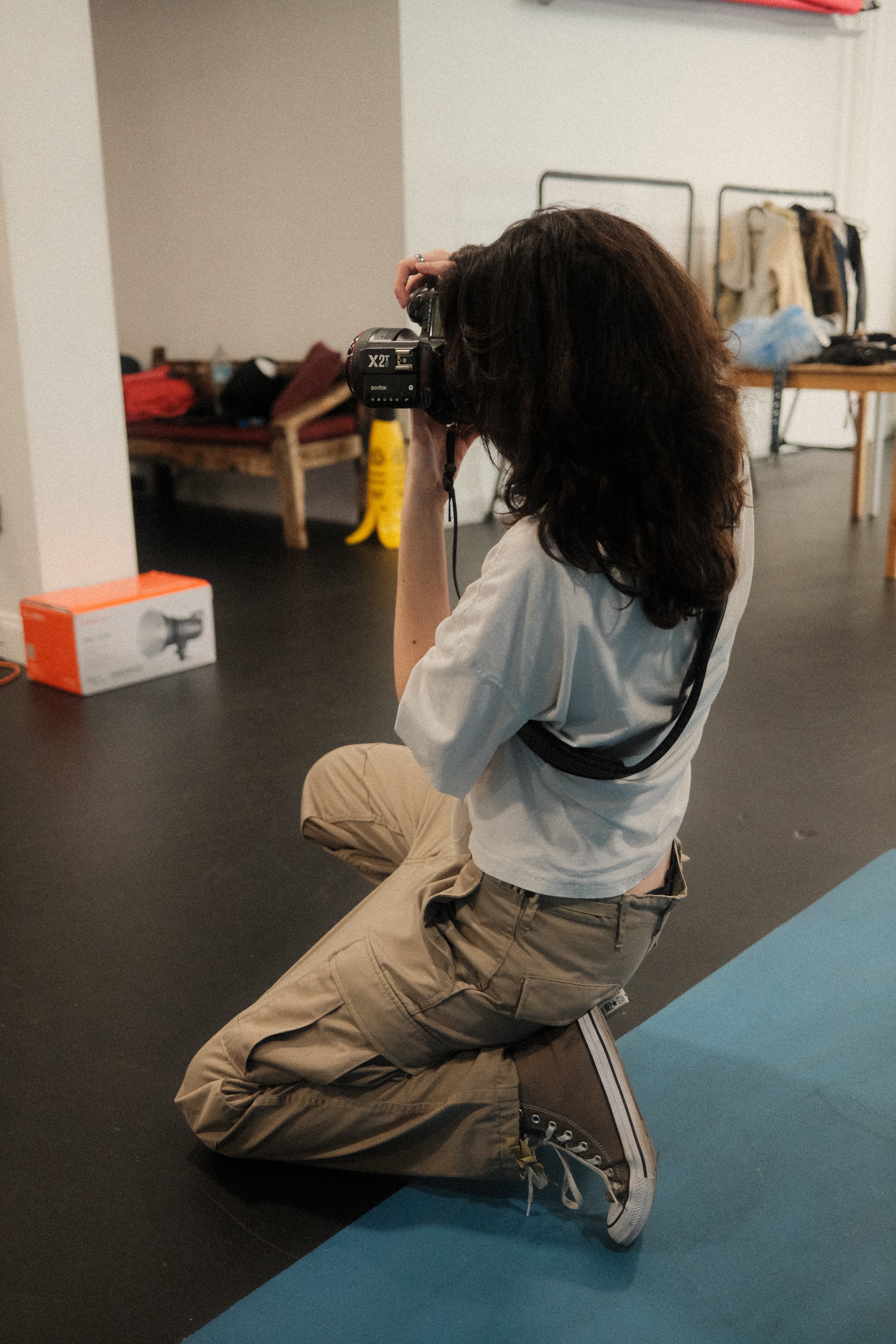 A person with curly brown hair is kneeling on a blue exercise mat, taking a photograph with a black camera.