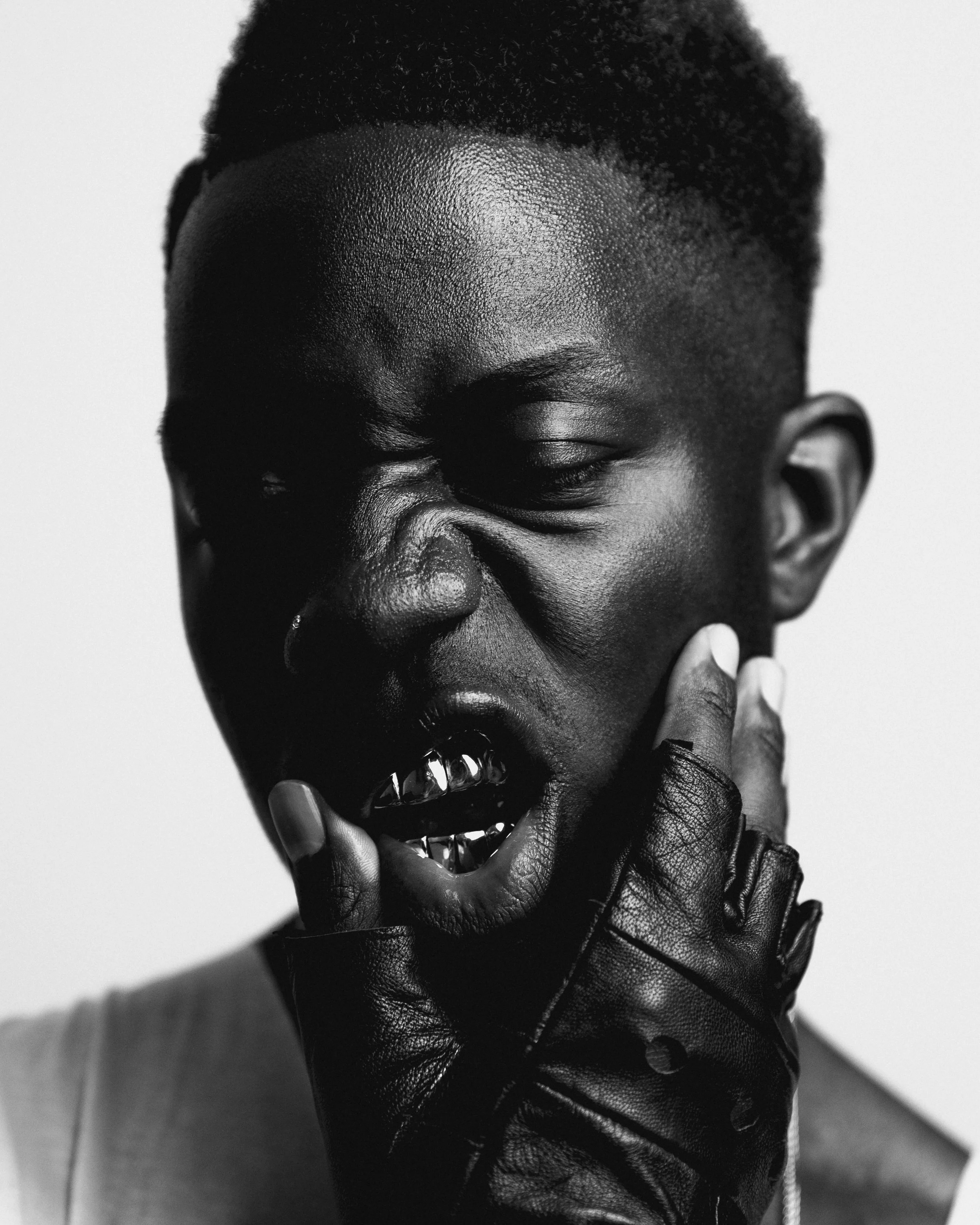 Black and white photo of a person with short hair and a nose piercing, scrunching their face and biting a finger with gold teeth in a leather glove.
