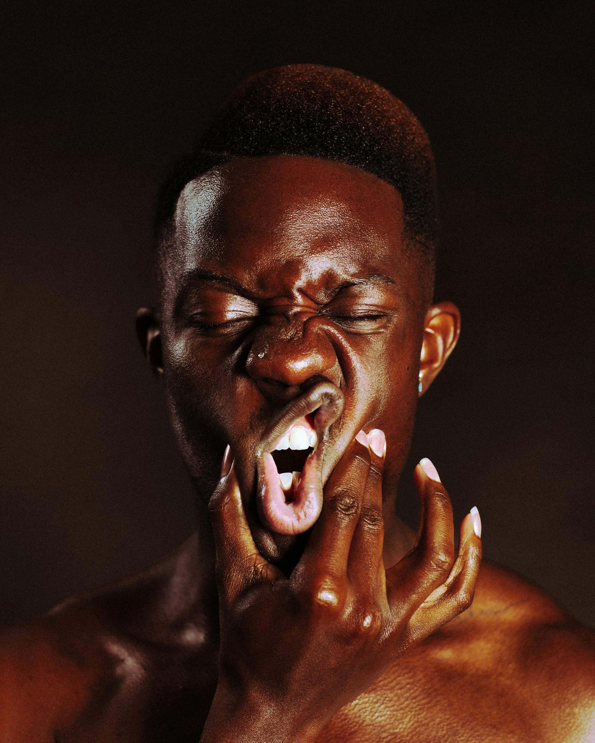 A shirtless man with dark skin and a "gumby" haircut, eyes closed, mouth open, hand grabbing his face, in front of a dark background.