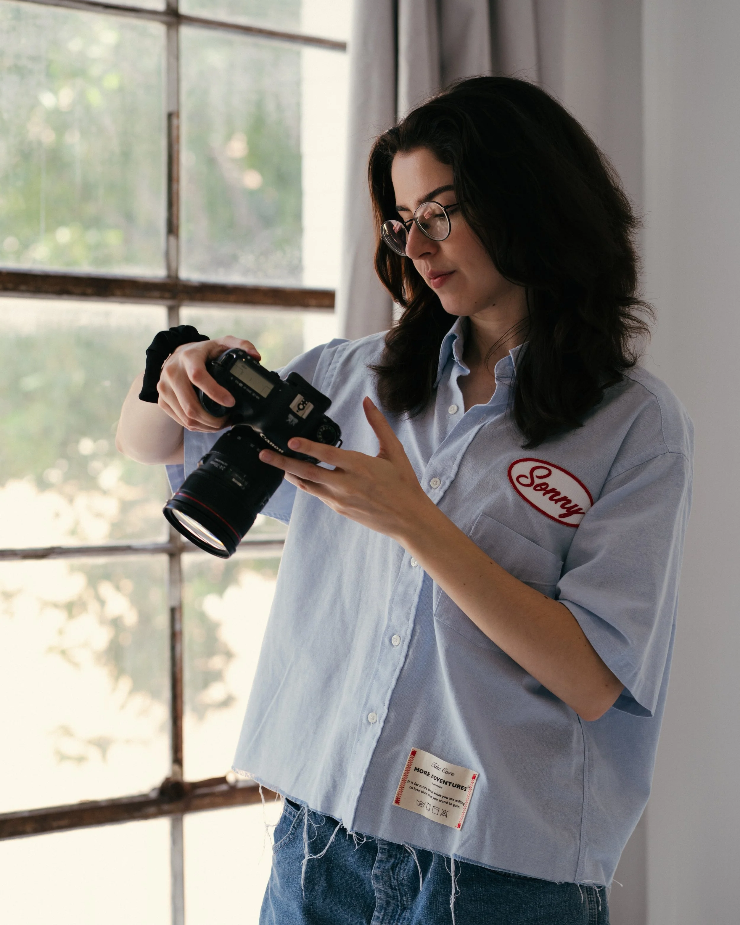 A woman with glasses and dark hair wearing a light blue button-up shirt looking at the screen of a camera she is holding.