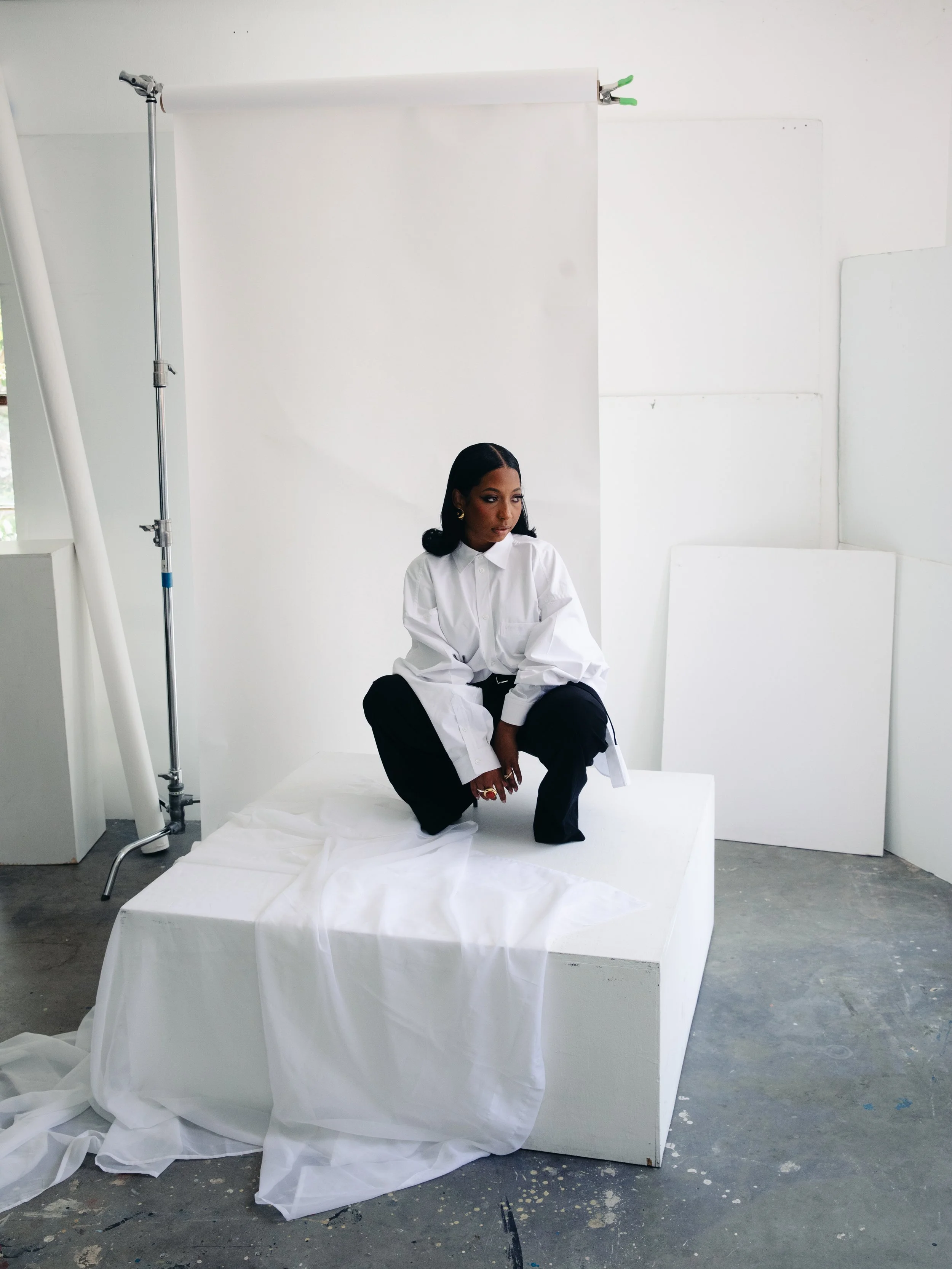 A woman dressed in a white button-up shirt and black dress pants crouches on a white box in a photography studio with a white backdrop and scattered white fabric.