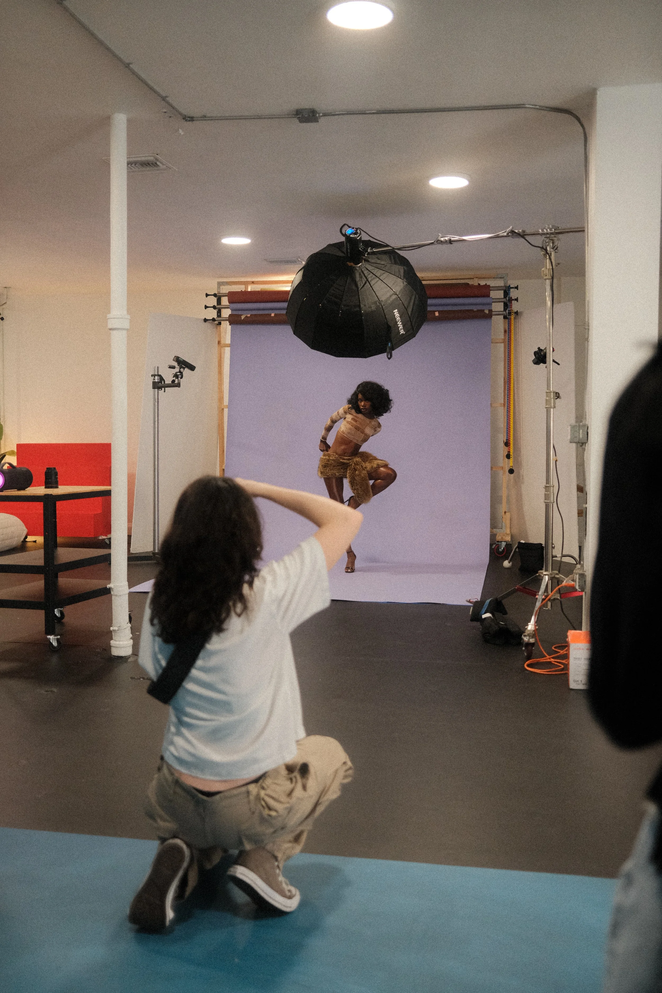 A photoshoot in a studio with a model dancing or posing, a photographer kneeling and taking pictures, and studio lighting and backdrop setup.