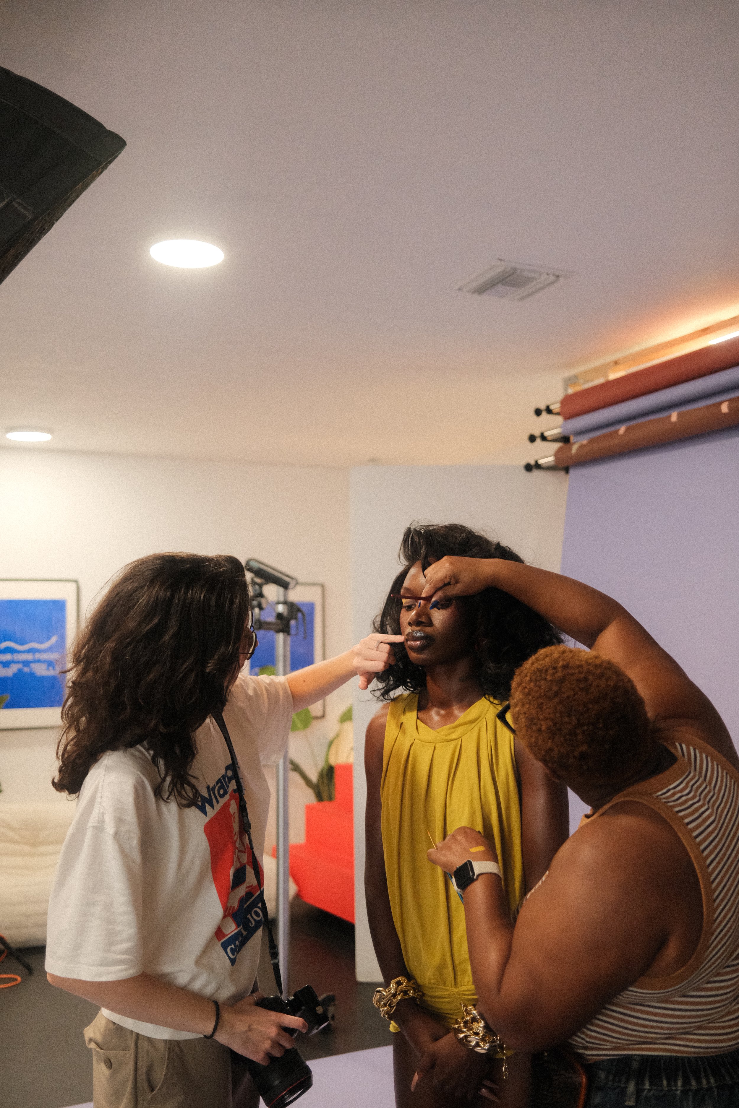 A woman in a yellow dress is having her makeup touched up by two makeup artists in a room with photography equipment and a purple backdrop.