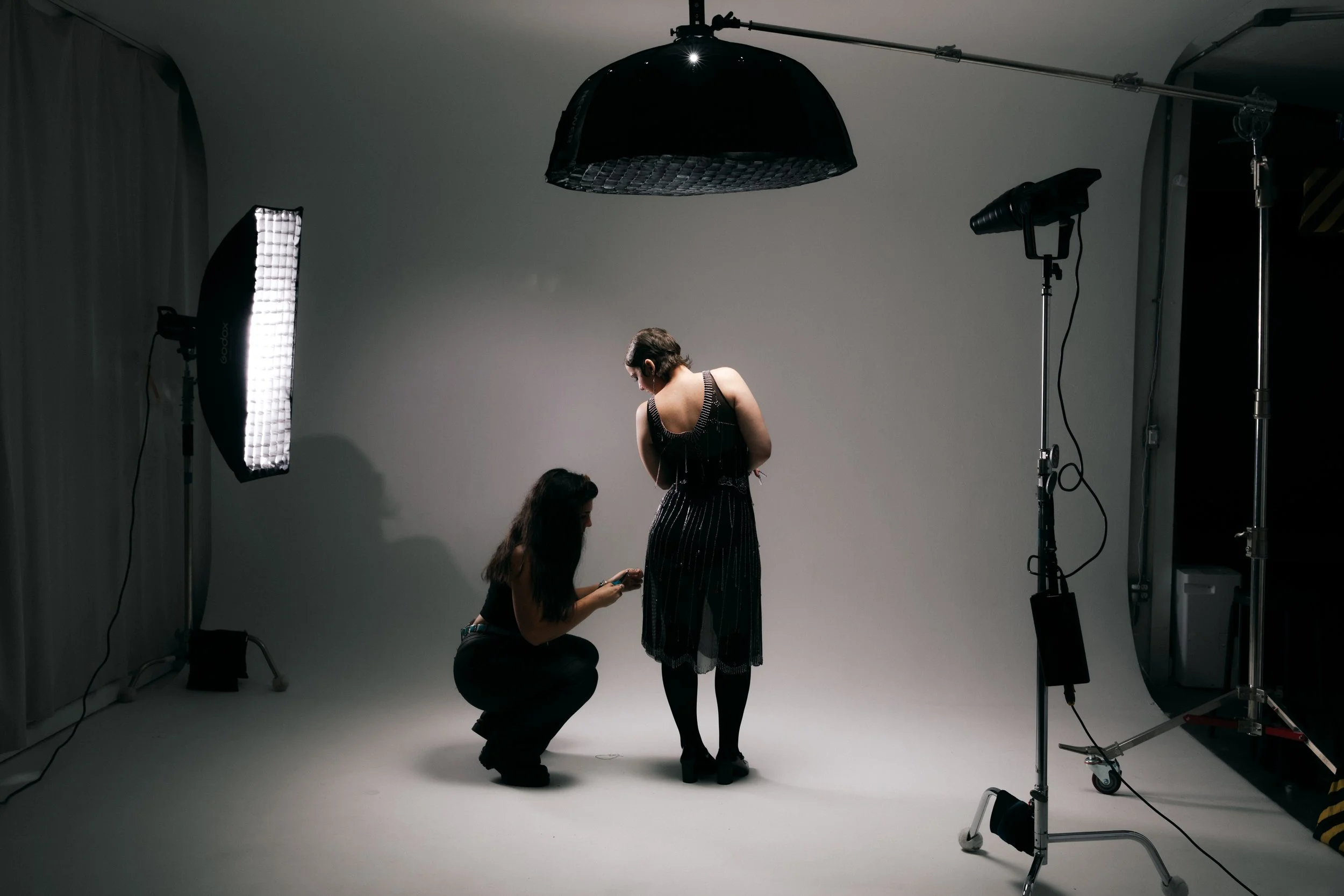Two women in a photography studio, one kneeling and the other standing, surrounded by professional lighting equipment and a plain backdrop.