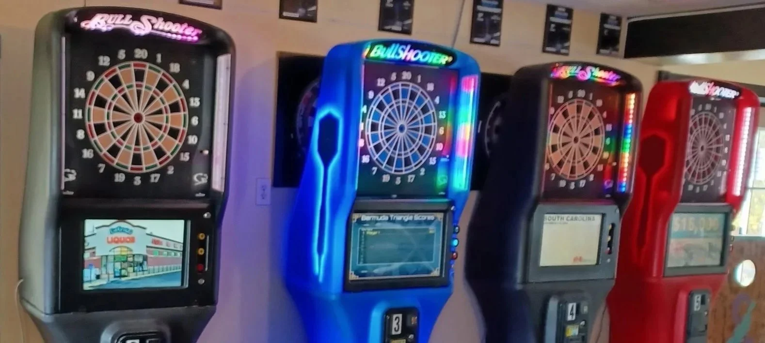 Four electronic dart machines lined up side by side in a game room. Each machine displays a digital dartboard on top and an electronic screen below.