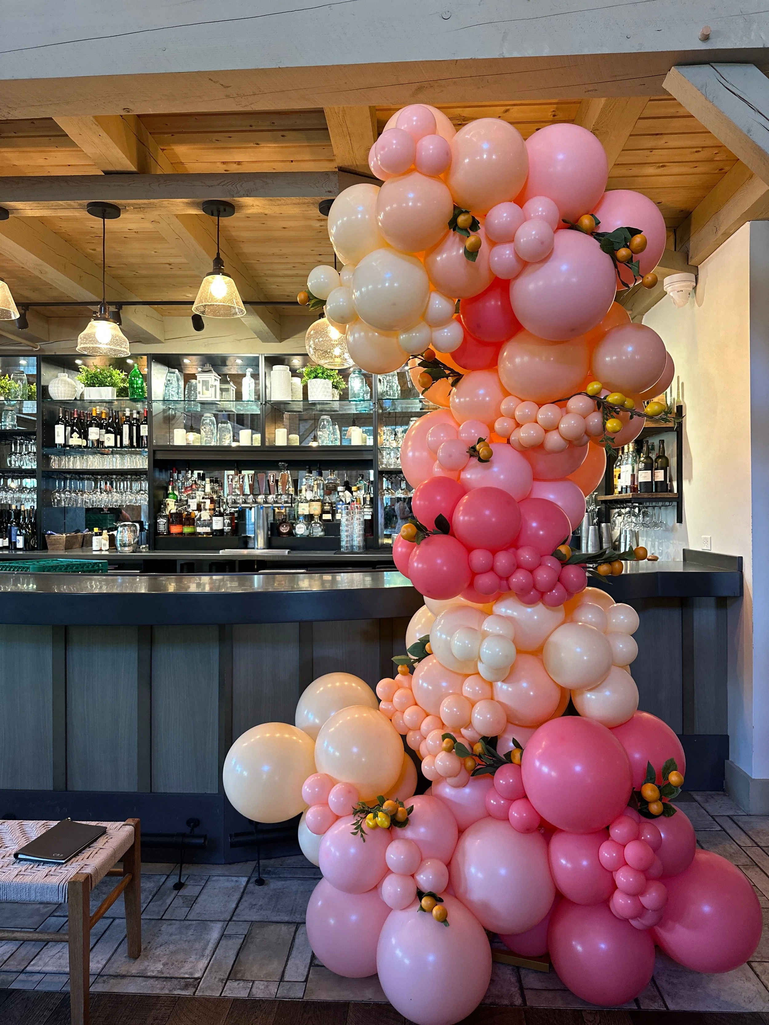 Decorative balloon arrangement in shades of pink, cream, and peach, with small orange fruits and green leaves, positioned next to a bar in a restaurant or cafe.