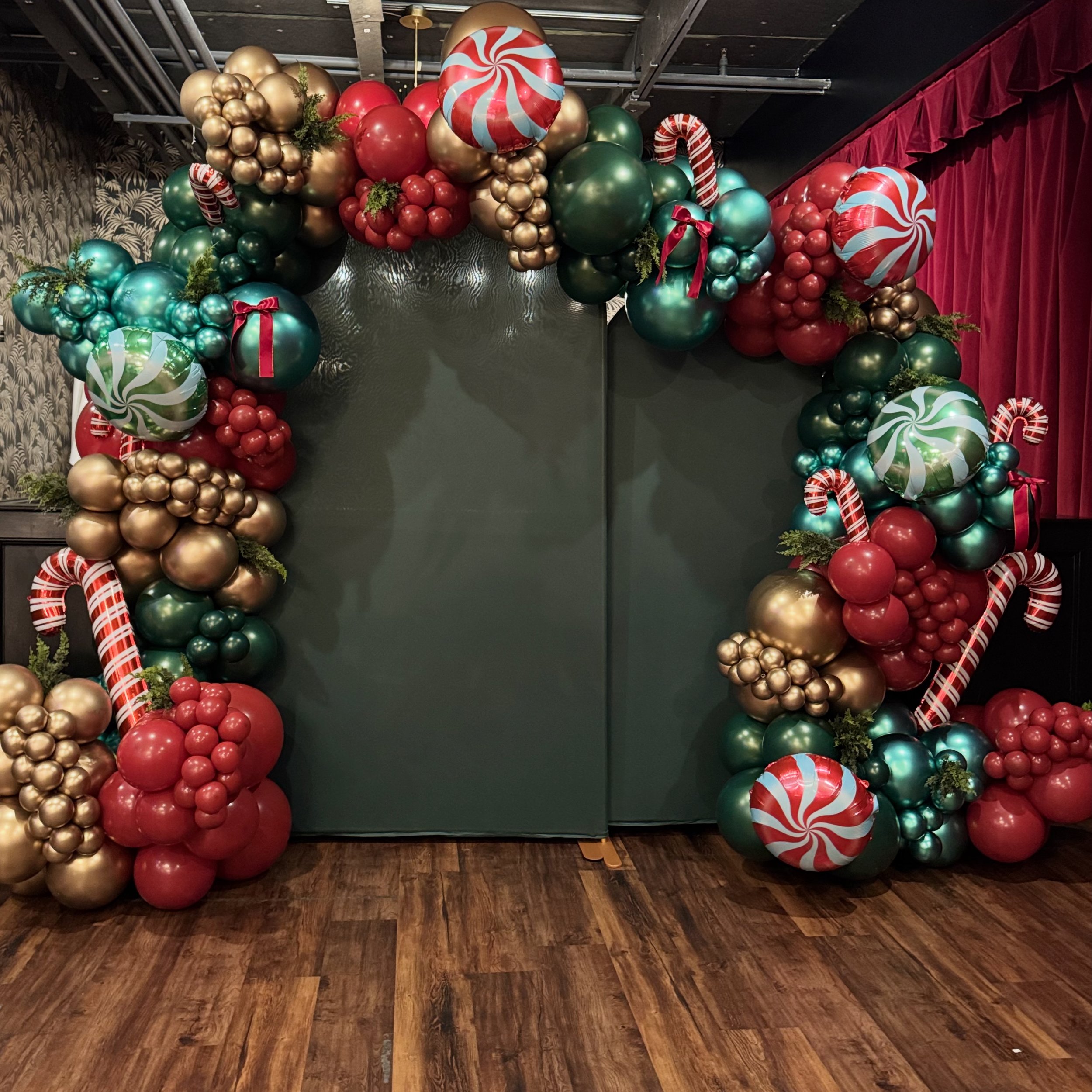 Holiday balloon decoration arch with red, green, gold, and candy-themed balloons, including peppermint swirls and candy canes, set against a dark green backdrop and red curtains.