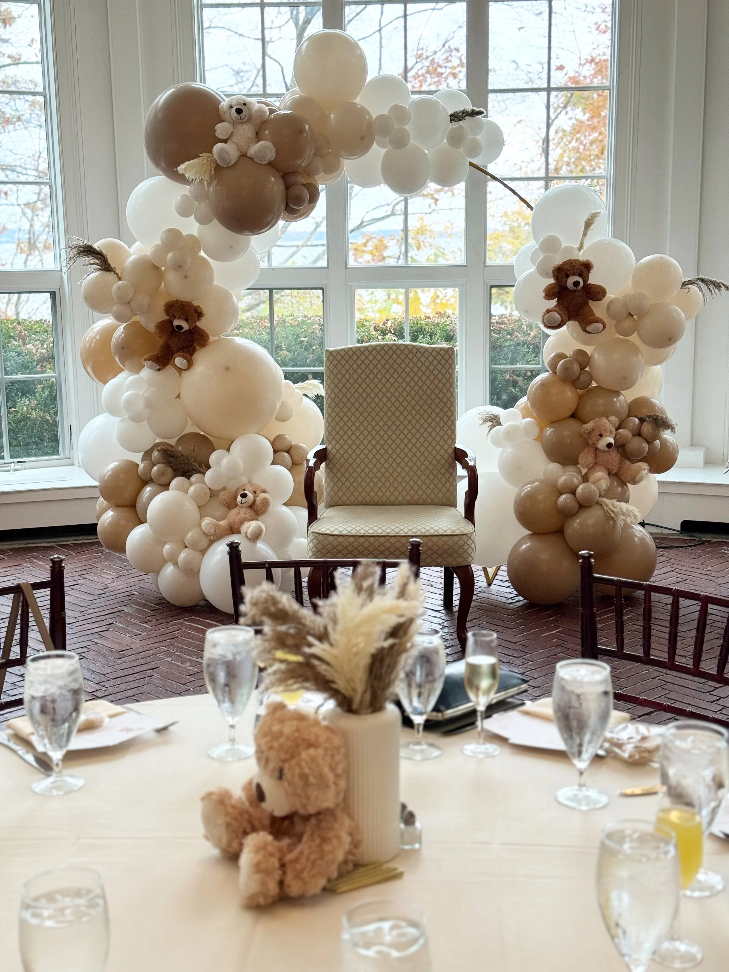 Decorative balloon arch with teddy bears and pampas grass, set up behind a vintage armchair in front of large windows, with a table arranged with glasses and a teddy bear centerpiece in a bright room