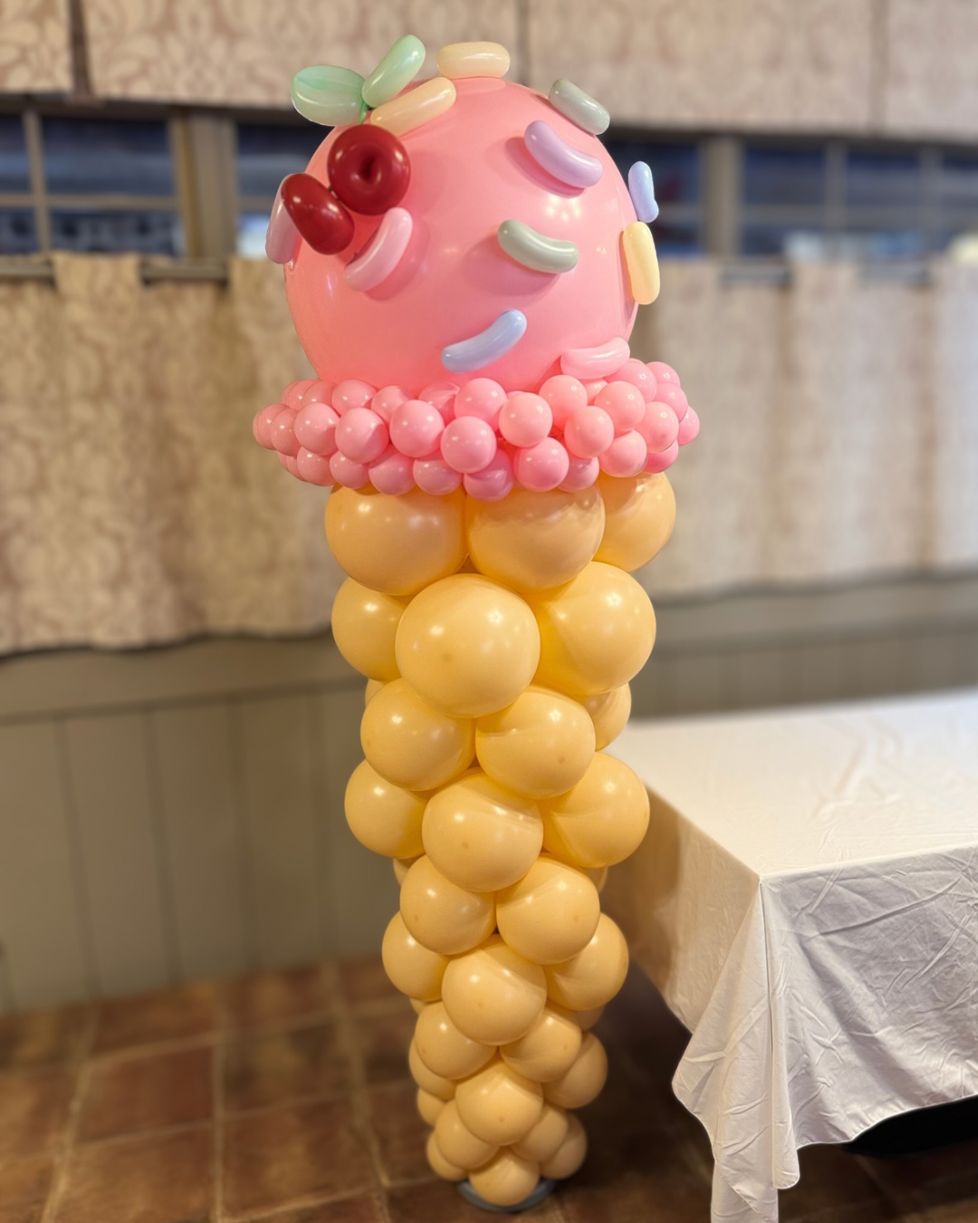 Balloon sculpture resembling an ice cream cone with a pink balloon scoop topped with colorful candy-topped balloons and a cone made of yellow balloons.