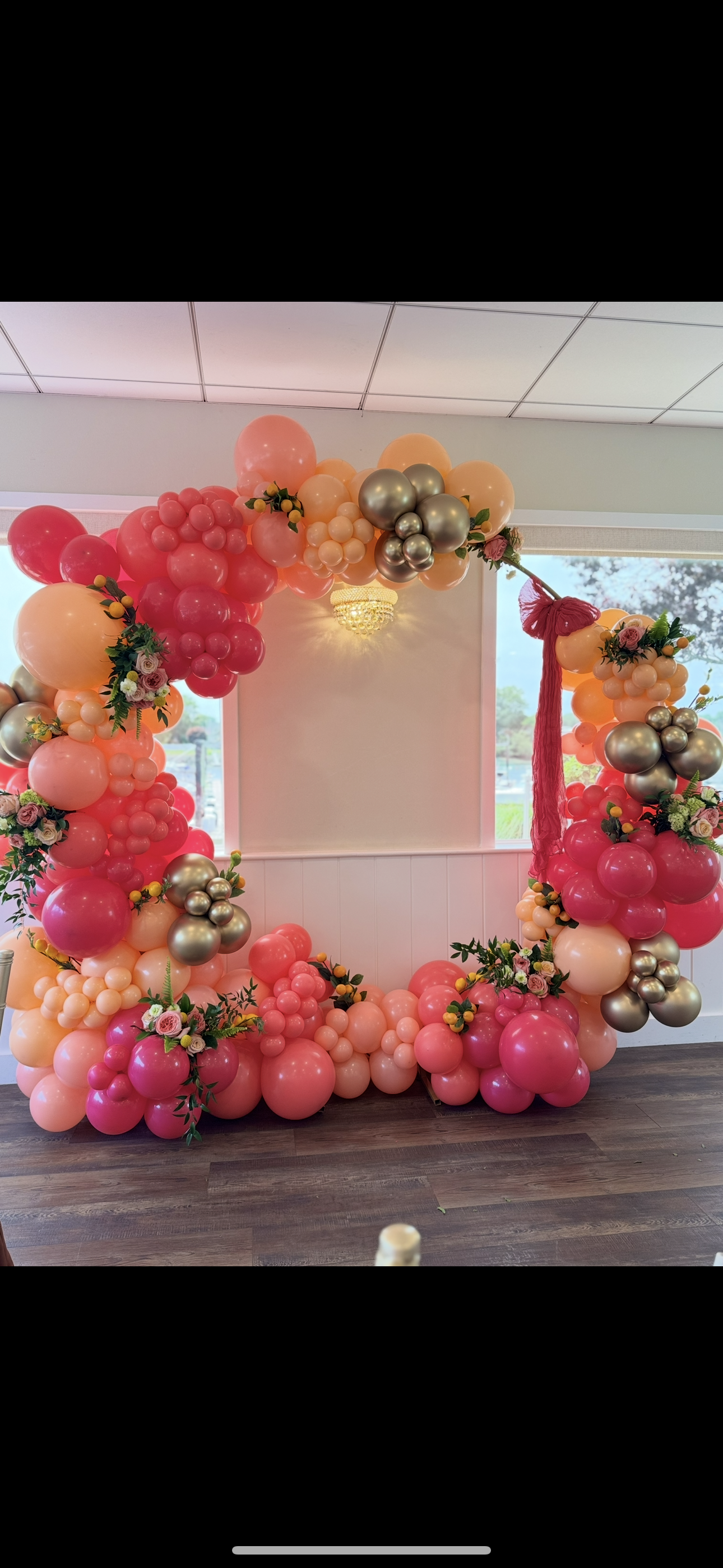 A colorful balloon arch with pink, peach, silver, and cream balloons, decorated with small bouquets of flowers and greenery, set in front of a window inside a room for a celebration or event.
