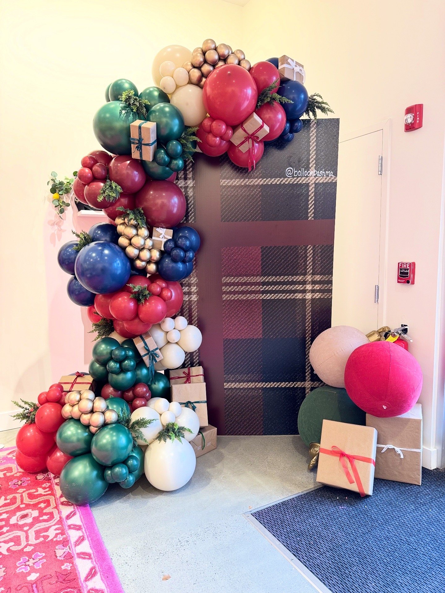 Holiday balloon display with red, blue, white, gold, and green balloons, decorated with gift boxes, greenery, and wrapped presents, located near a plaid-patterned wall and an exit door.