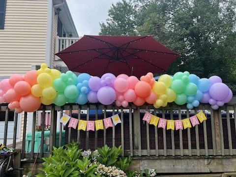 Colorful balloon arch and "Happy Birthday" banner on a deck railing with an umbrella.