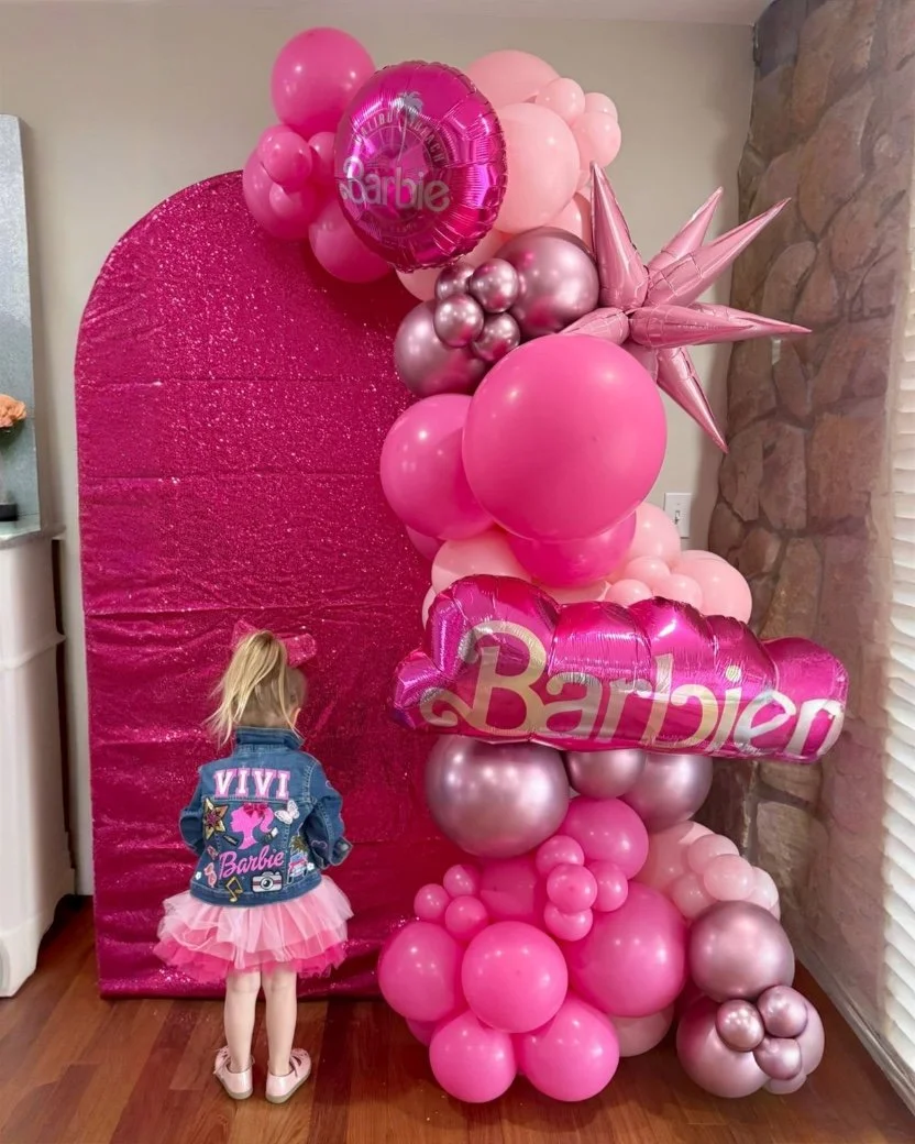 A young girl in a denim jacket with a pink tutu skirt stands near a pink balloon display with a sparkling pink backdrop. The balloons are various shades of pink and metallic, forming a large decorative arrangement with a shiny pink "Barbie" balloon a