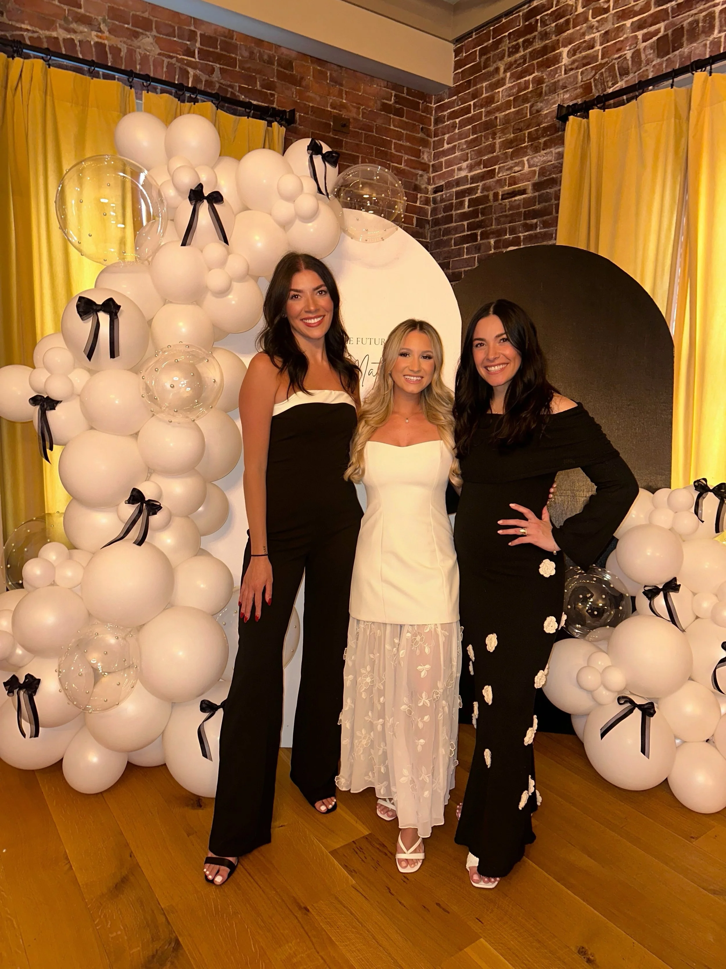 Three women dressed in formal attire standing in front of a balloon arch with white balloons and black ribbons, against a brick wall with yellow curtains.