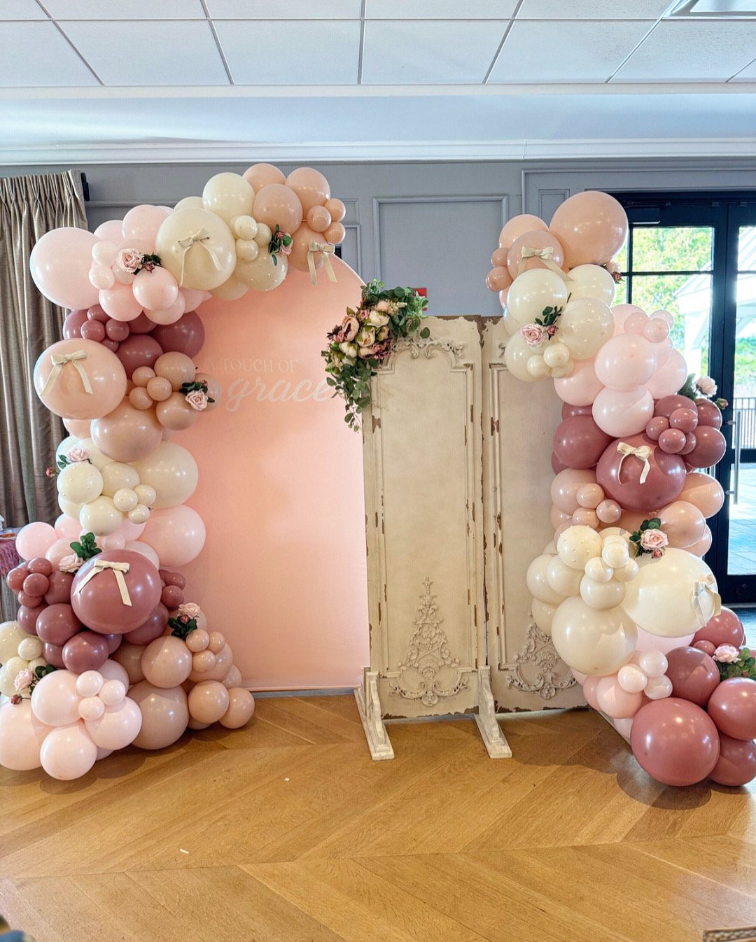 Decorative balloon arch with pink, white, and blush balloons, small flowers, and ribbon accents, surrounding a pink backdrop with the text 'touch of grace', and a vintage white wooden screen in a room with wooden flooring, curtains, and a glass door.
