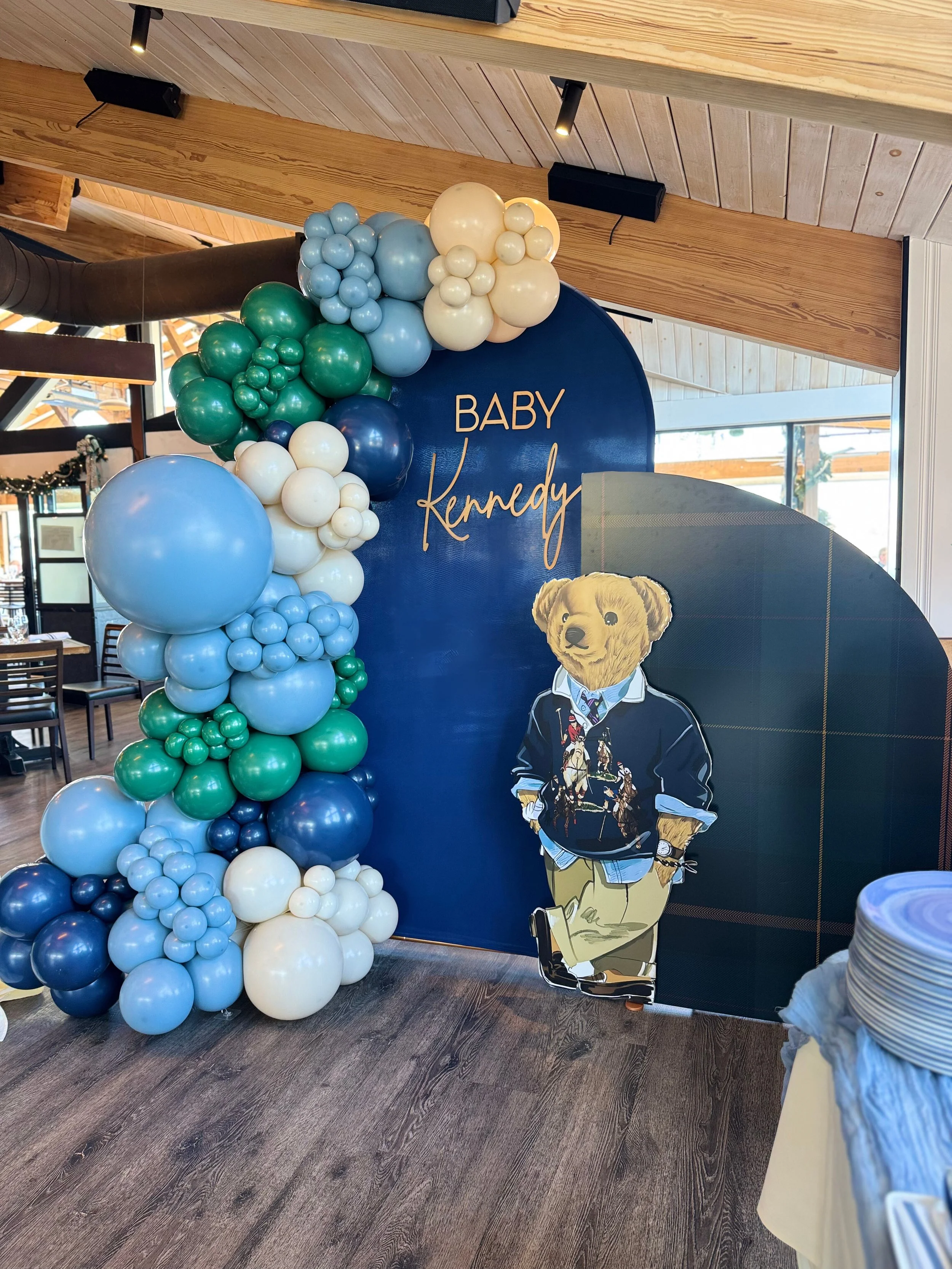 Decorative display for a baby shower or celebration with a blue backdrop that has the words "BABY Kennedy" and a large teddy bear cutout. There is an arrangement of blue, white, and green balloons on the left side of the backdrop.