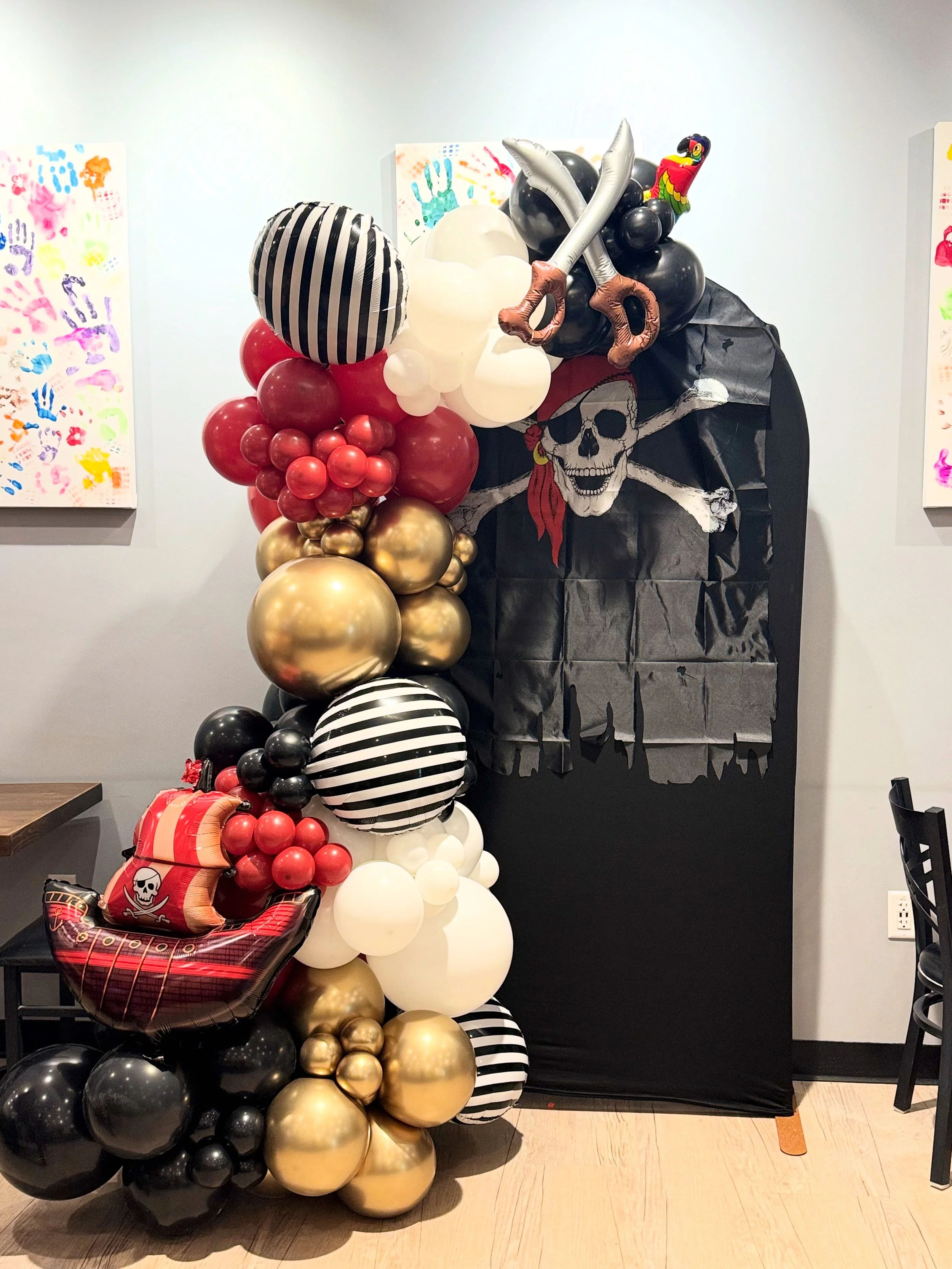 Pirate-themed balloon arch with black, white, red, and gold balloons, featuring a balloon pirate ship and a black backdrop with a skull and crossbones.