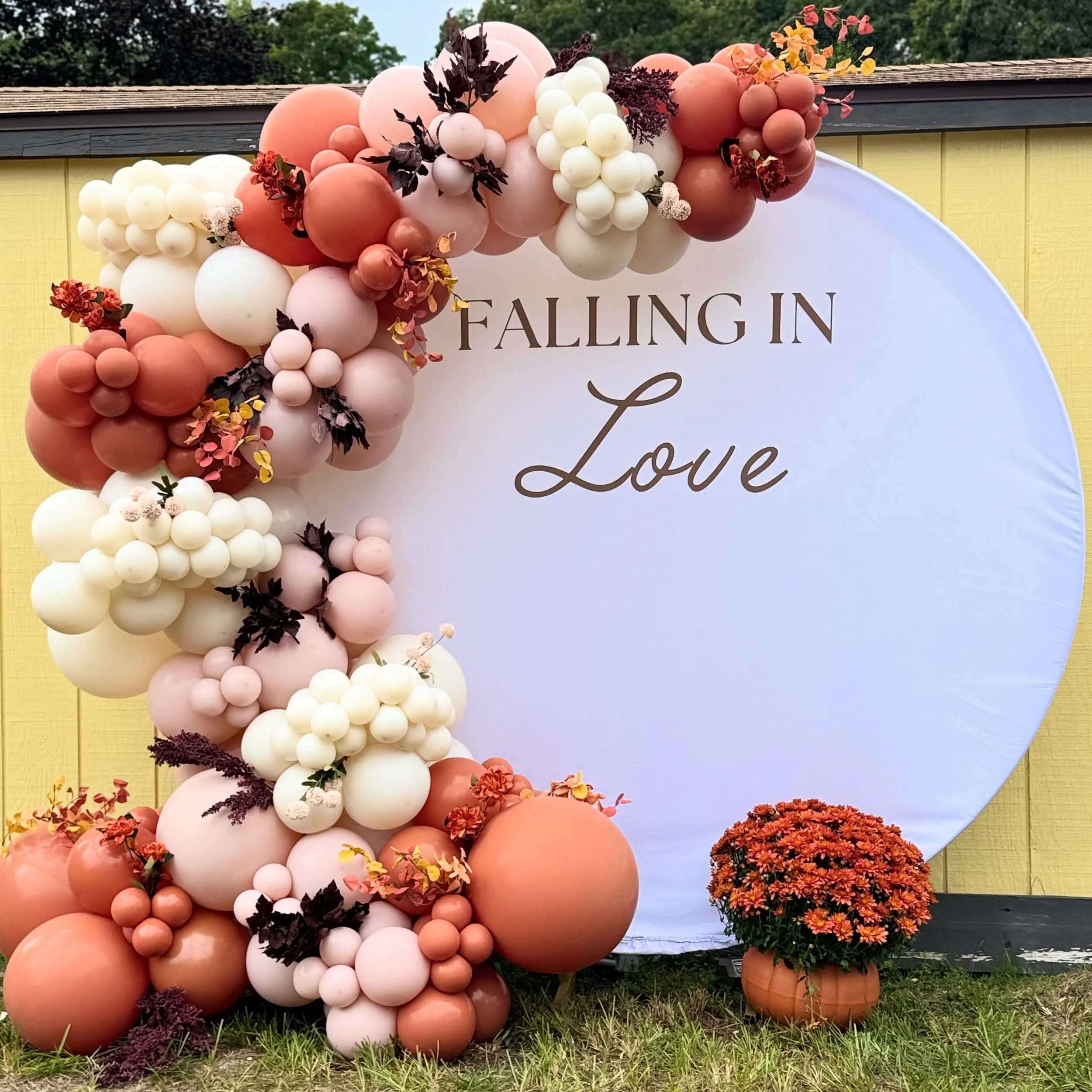 Decorative balloon arrangement with pink, white, peach, and maroon balloons around a white circular backdrop that reads 'Falling in Love'. There is a potted orange flowering plant at the base.