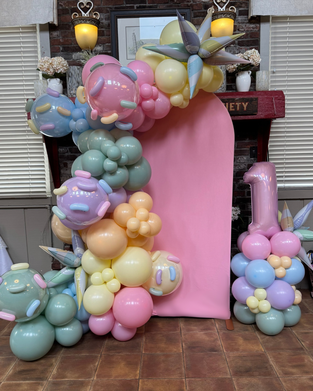 Balloon arrangement with pastel-colored balloons and a large pink number 1 balloon, decorated around a pink backdrop in a festive setting.