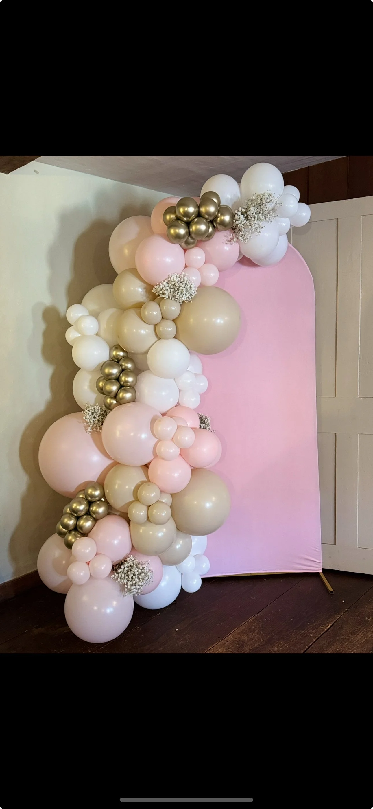 Decorative balloon arrangement with pink, white, gold, and cream balloons, accented with small white flowers, placed against a pink backdrop