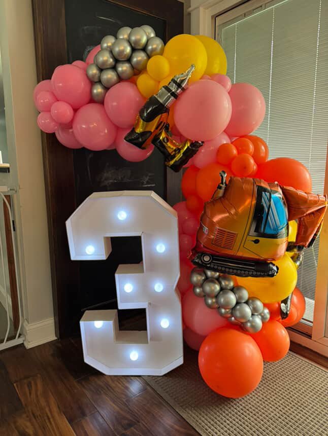 Large white illuminated number three surrounded by pink, orange, yellow, and silver balloons, with construction vehicle and champagne bottle balloon decorations.