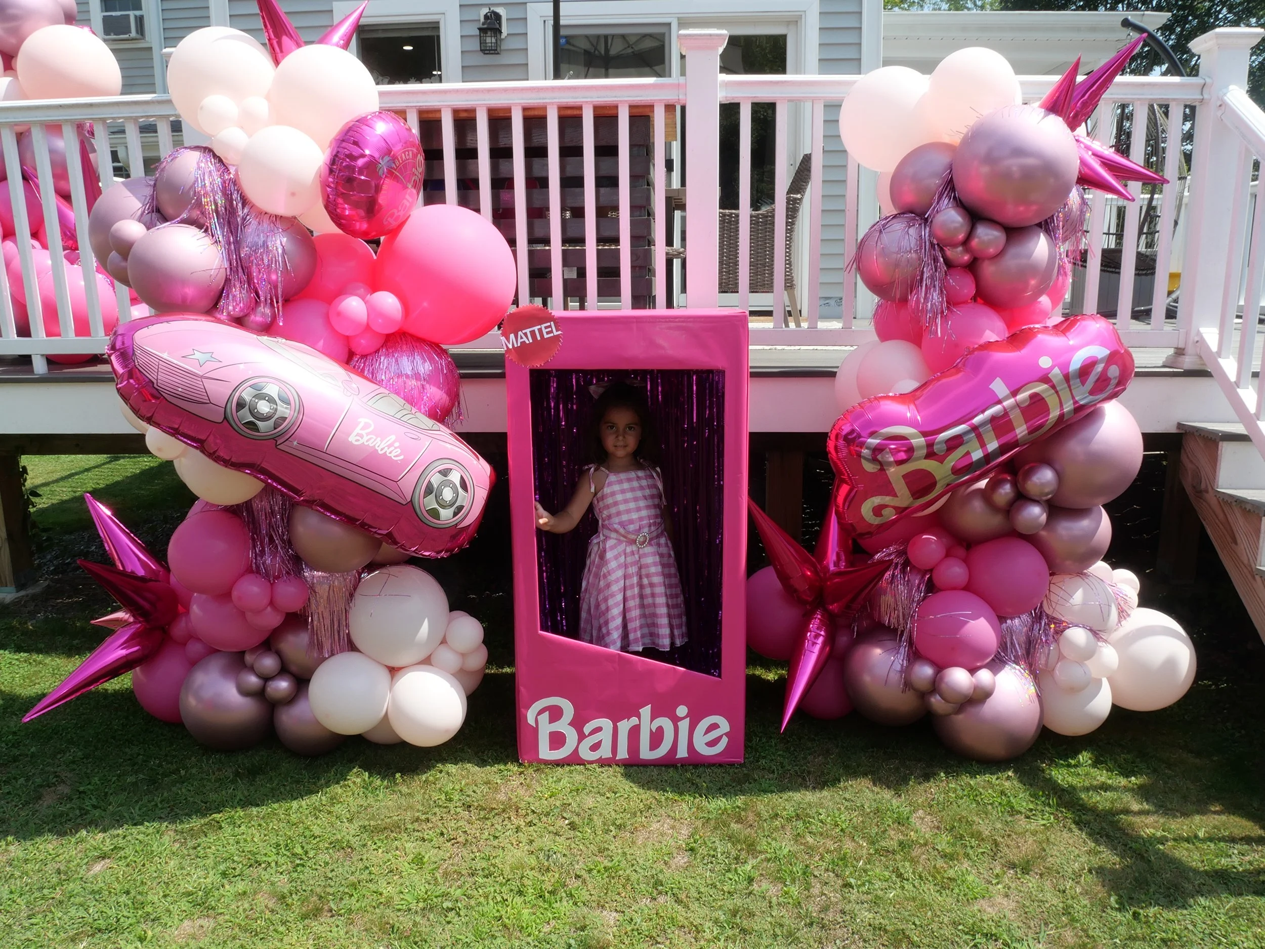 A young girl in a pink and white checkered dress standing behind a pink Barbie-themed photo booth, decorated with pink, white, and silver balloons, including large Star Wars-themed balloons, and metallic pink and silver accents, set up outdoors on a 