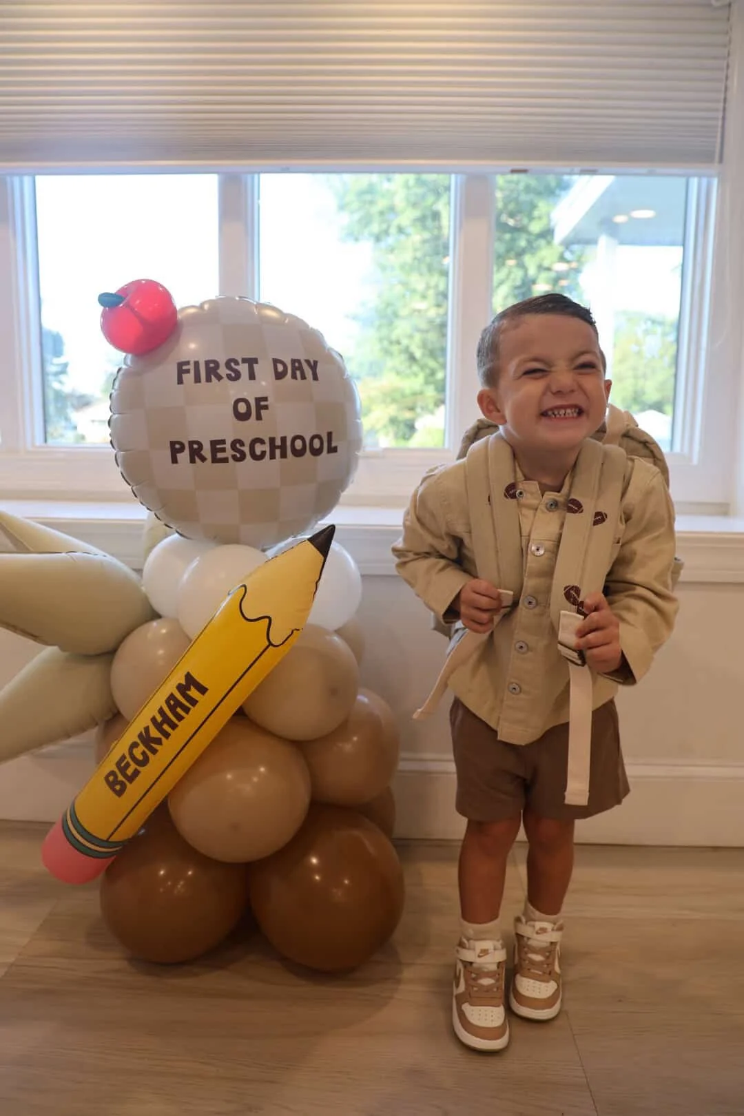 A young boy standing indoors in front of a window, smiling with his teeth showing, wearing a beige shirt with a backpack. Next to him is a balloon decoration with a round balloon reading 'First Day of Preschool,' a bunch of smaller white balloons, a 