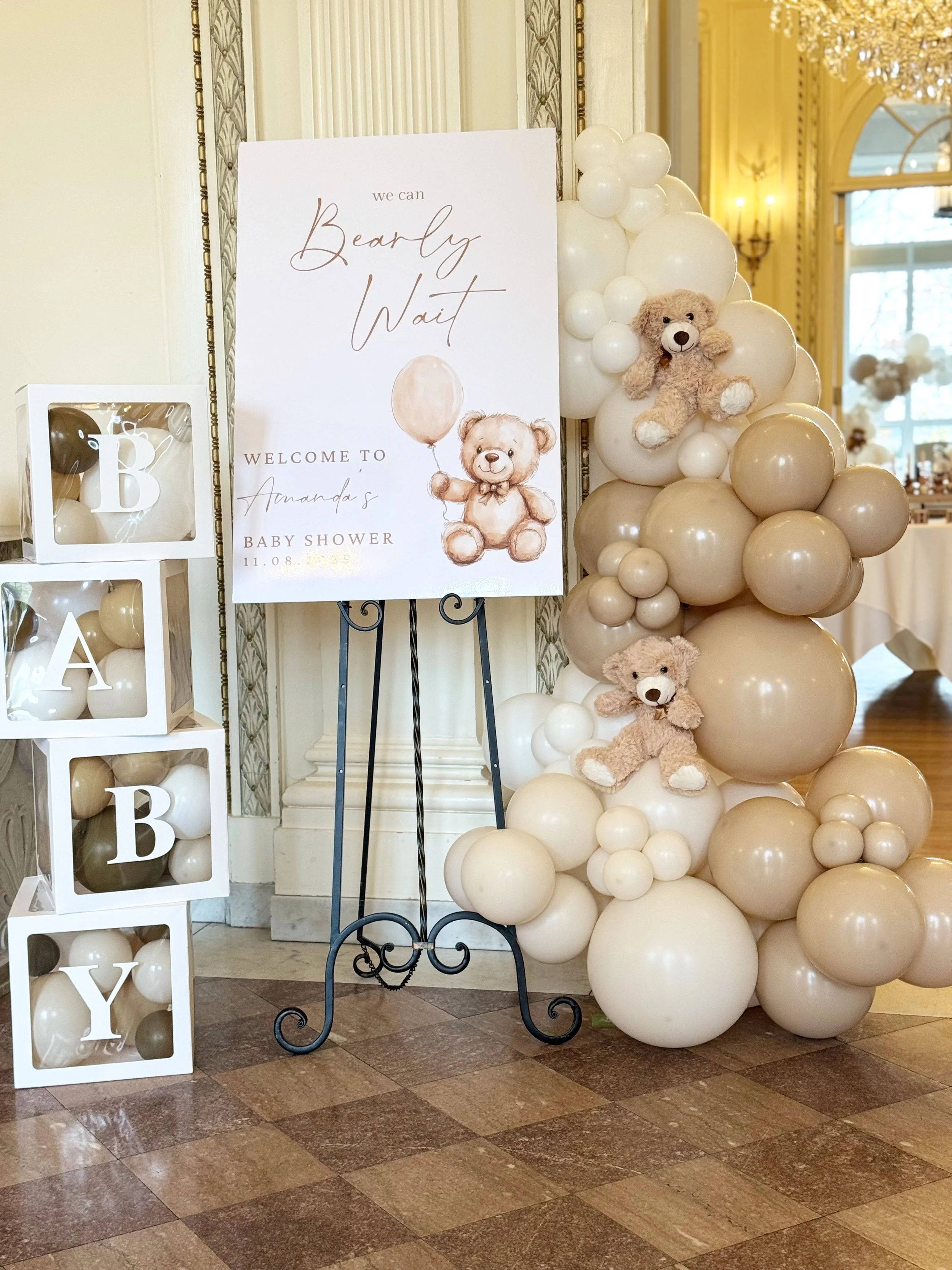 Decorations for a baby shower, featuring a balloon backdrop in cream, white, and beige with teddy bears, a welcome sign on an easel, and a cube display spelling 'BABY' with matching balloons.