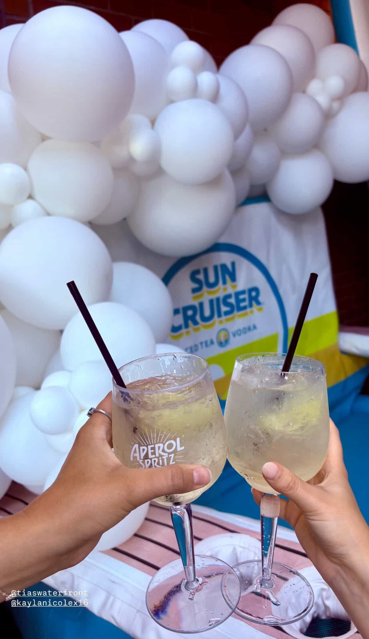 Two hands holding cocktail glasses with lemon slices, raised in a toast, in front of a backdrop of white balloons and a Sun Cruiser banner, at an outdoor event.