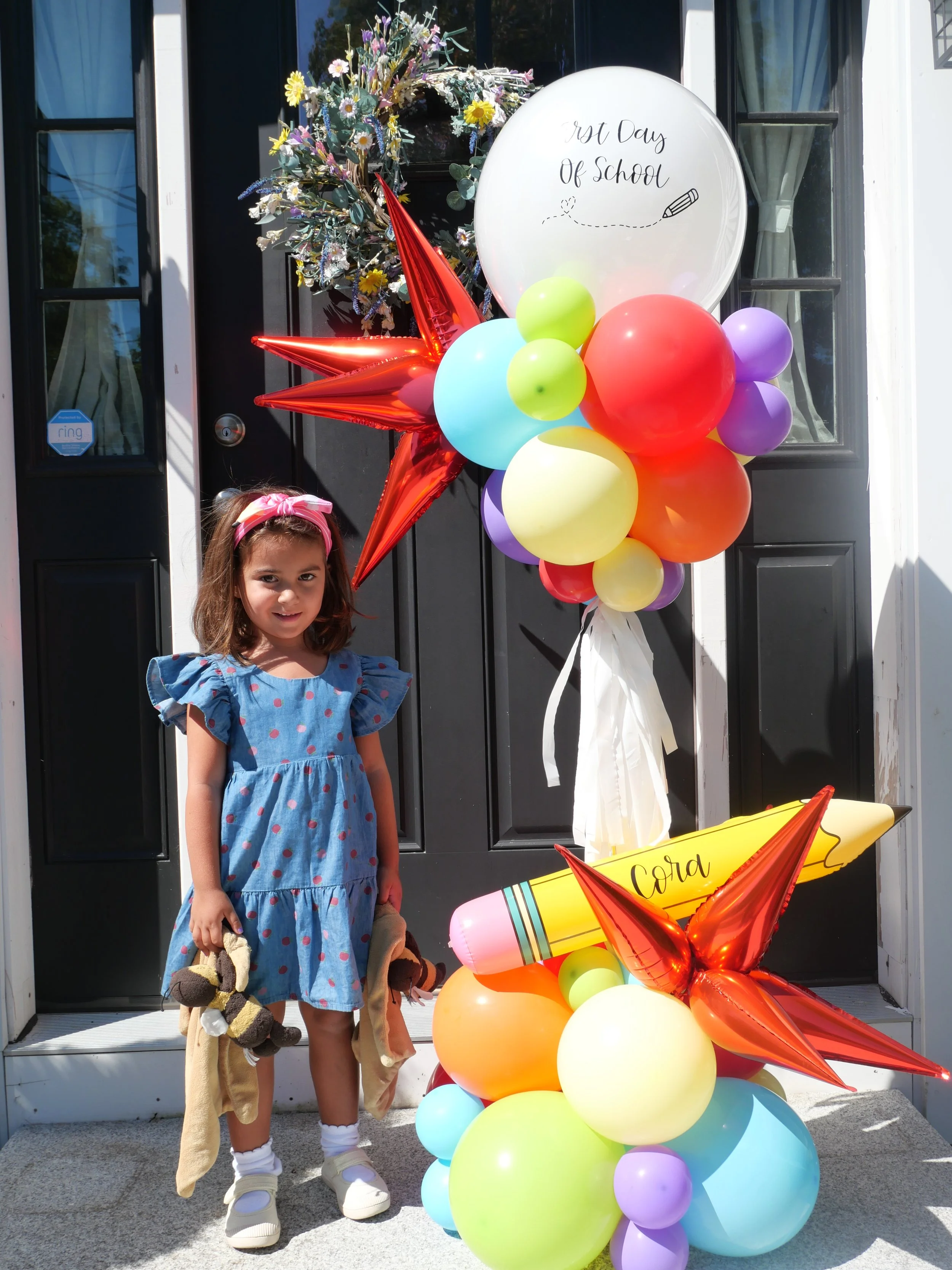 A young girl standing in front of a decorated front door with balloons, a large red star-shaped balloon, and a sign that reads '1st Day of School.' The girl is holding a plush toy and a small bag, wearing a blue dress with pink polka dots and a pink 