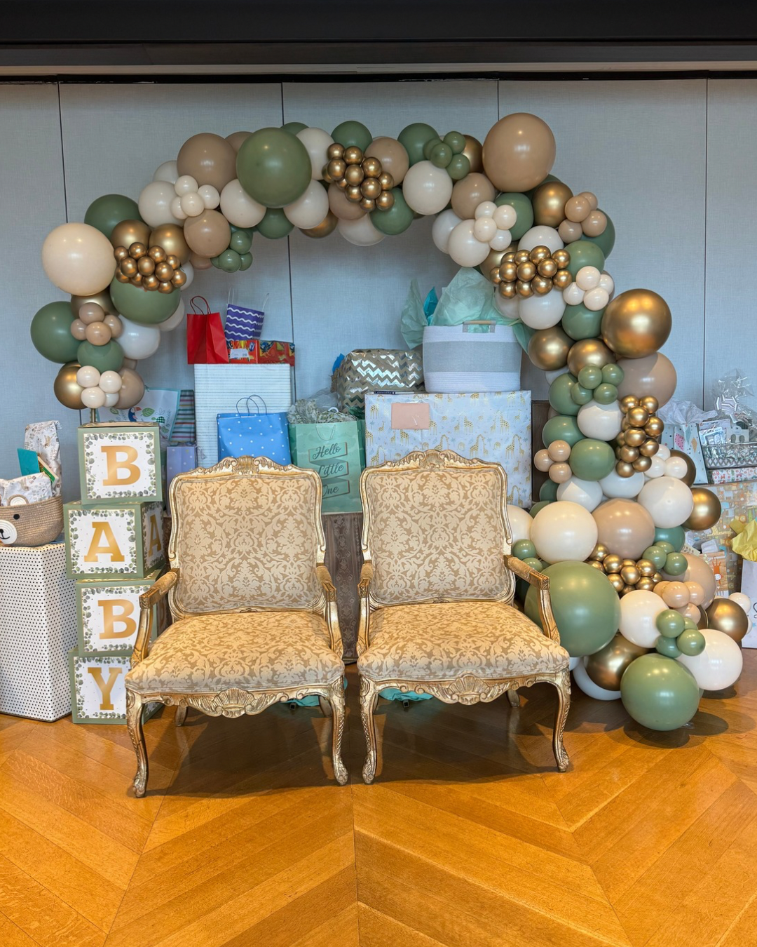 A decorative balloon arch with gold, white, and green balloons surrounds two vintage upholstered chairs with gold patterned fabric. There are wrapped gifts and presents behind the chairs.