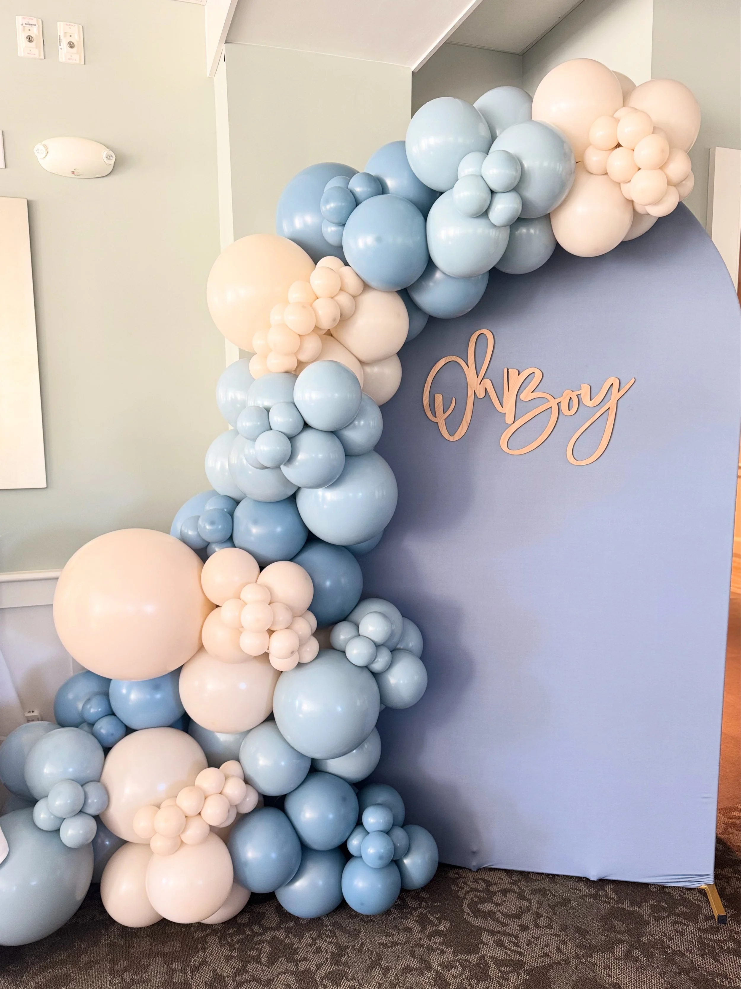 Arched balloon decoration in pastel blue, white, and cream, with a sign that reads 'Oh Boy' attached to a fabric backdrop, indicating a baby shower for a boy.