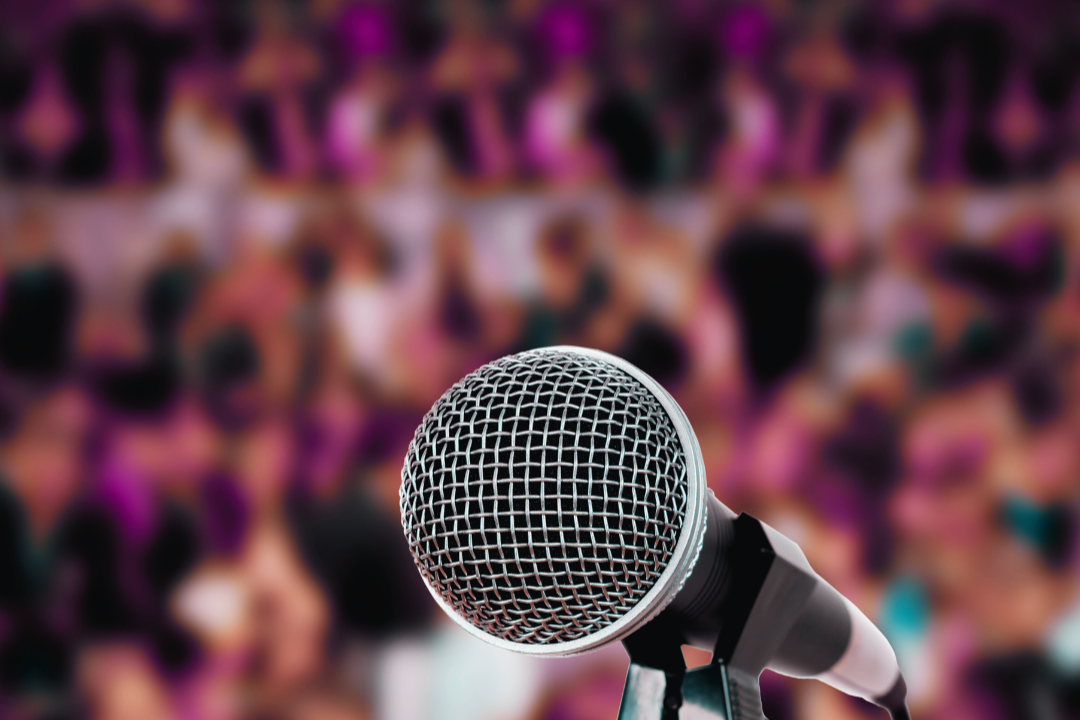 A large silver mic in the foreground. In the background is a blurred out audience.