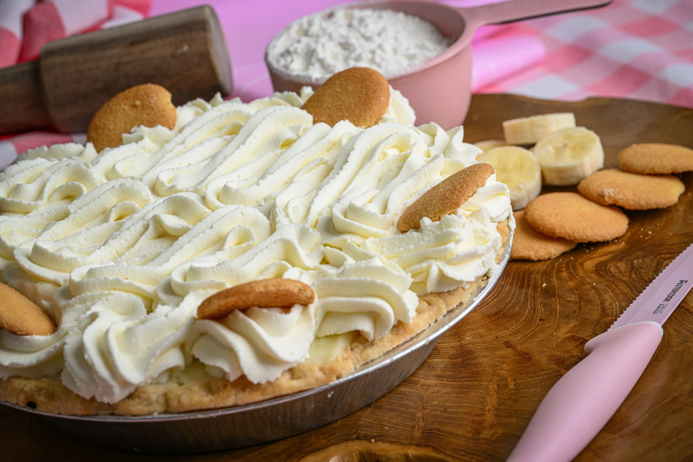 Banana cream pie topped with whipped cream, banana slices, and cookies, on a wooden surface with a pink rolling pin and baking ingredients in the background.