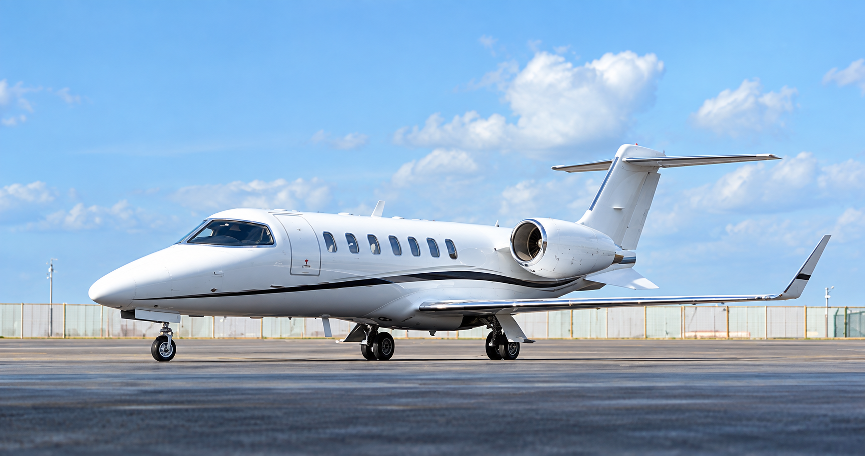 Bombardier Learjet 40 private jet parked on an airport apron, shown in a side exterior view under a bright blue sky with scattered clouds.