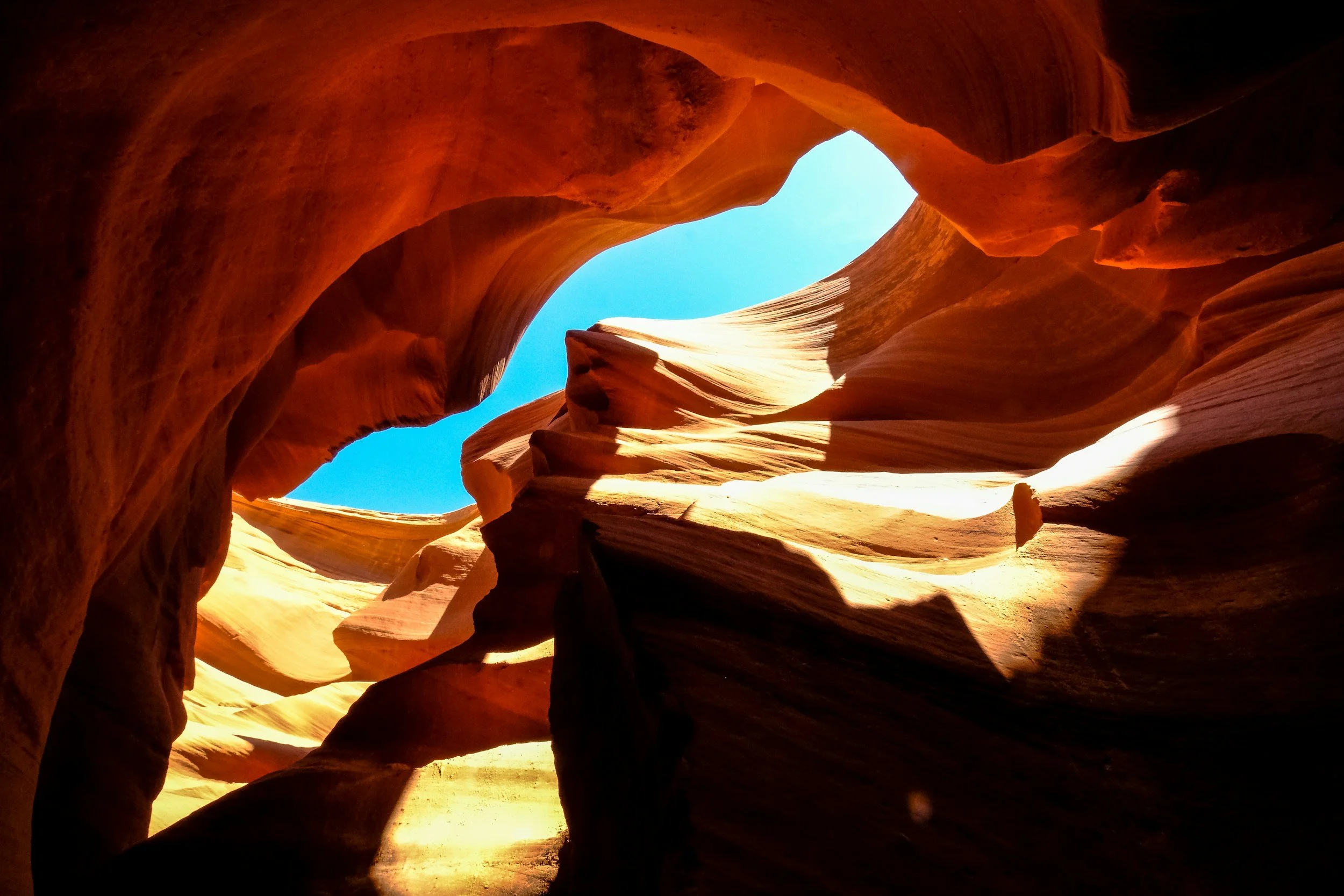View from inside a narrow slot canyon looking up at a blue sky through an opening of rock formations with orange and tan hues.
