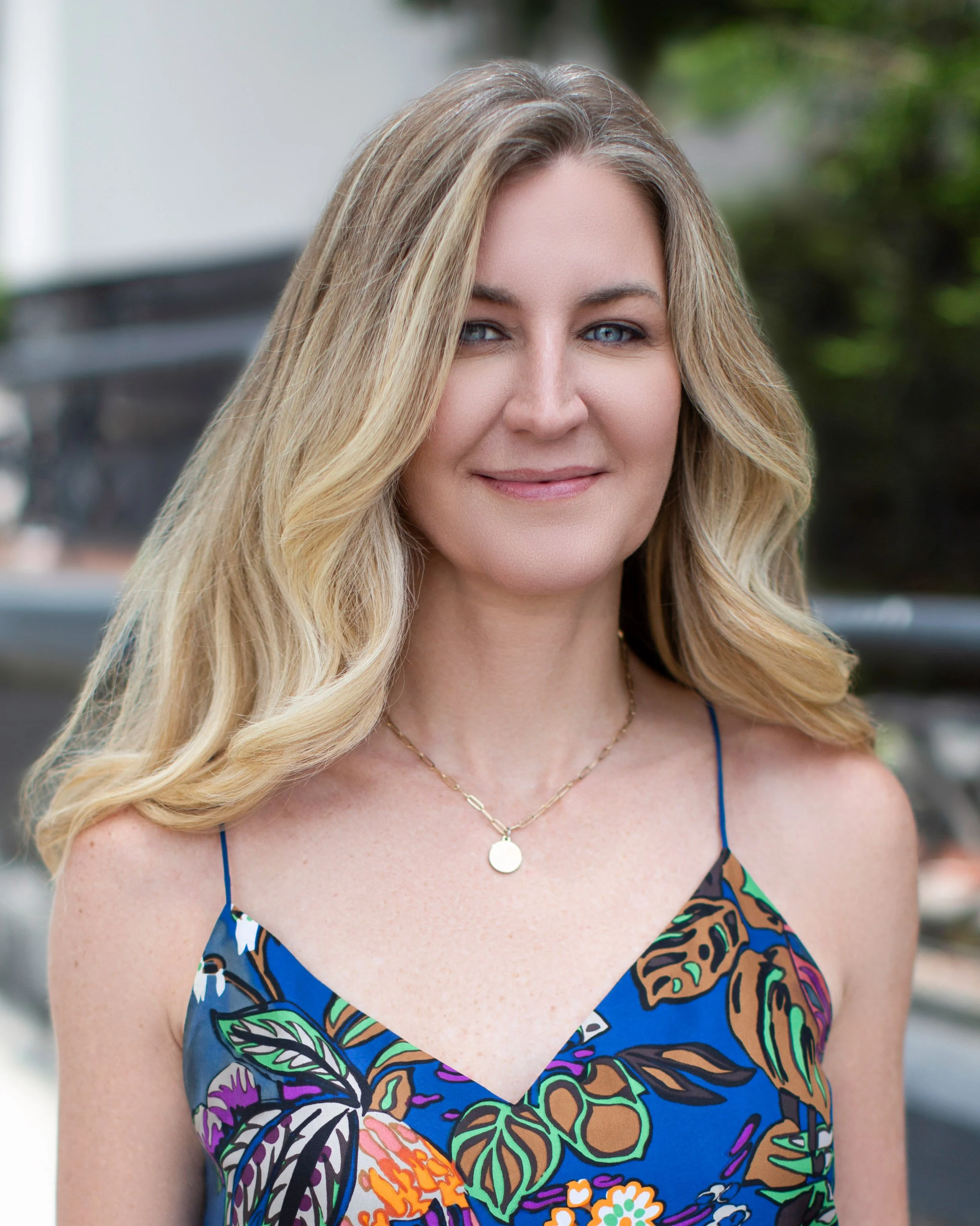 A woman with long blonde hair wearing a blue floral dress and a gold necklace, standing outdoors.