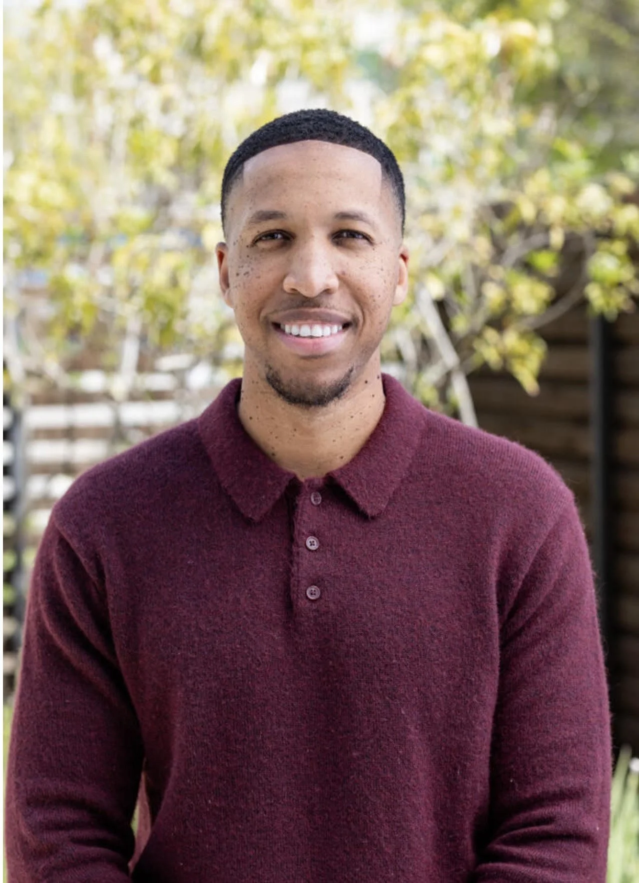 A smiling man with short dark hair and a goatee wearing a maroon sweater outdoors with green foliage and wooden fence in the background.