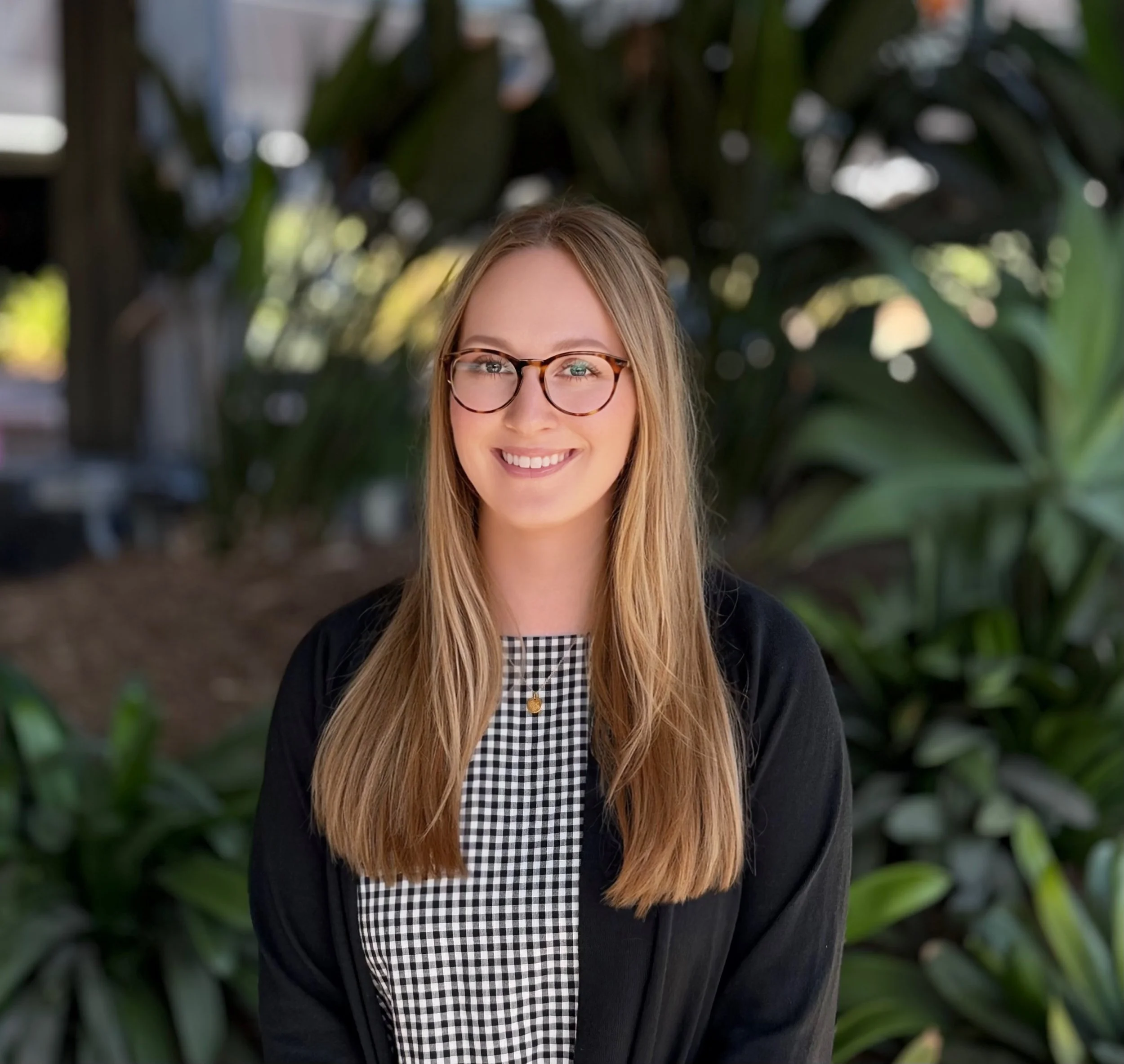 A young woman with long blonde hair, glasses, and a black and white checkered shirt smiling outdoors with green foliage in the background.